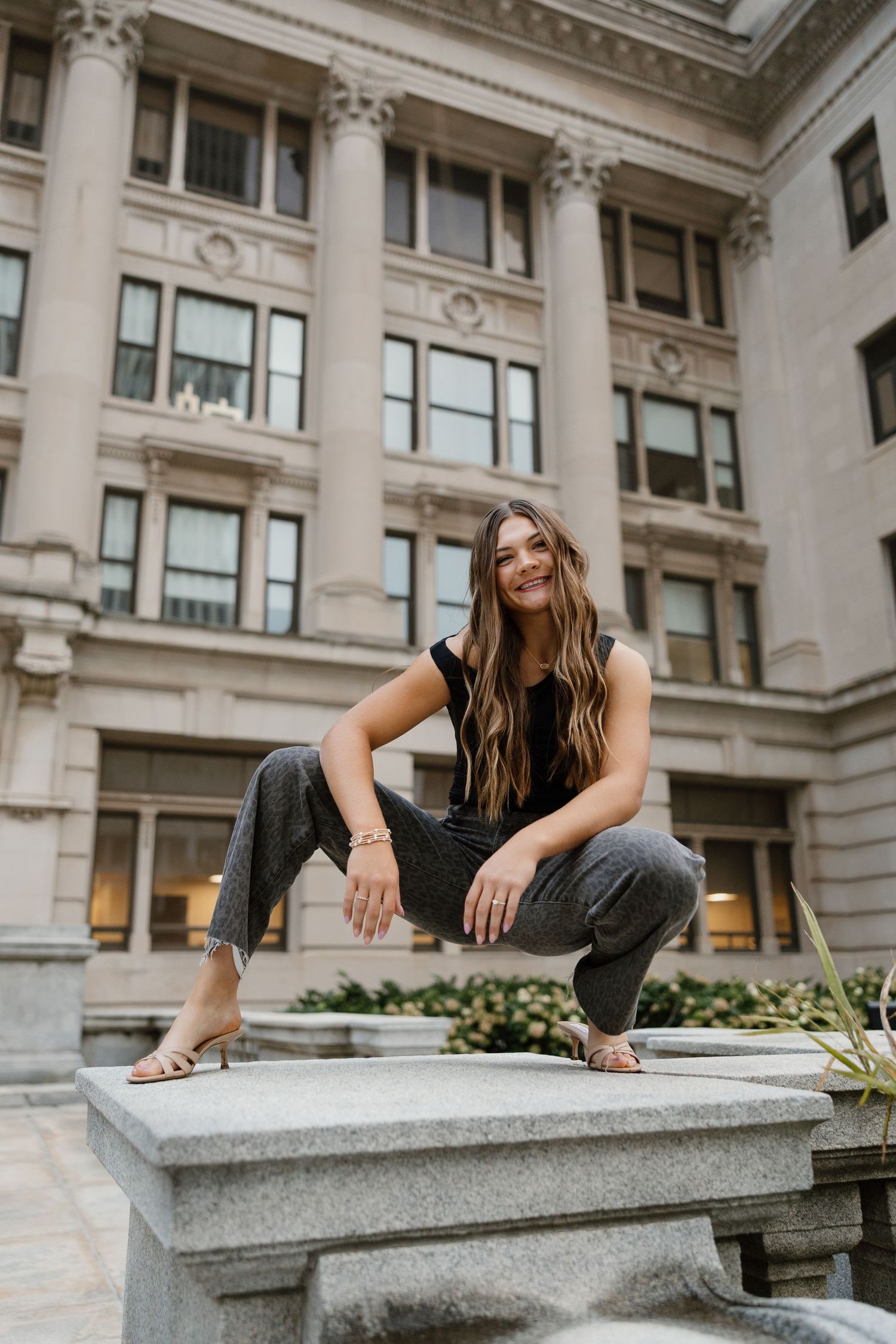 A person in casual attire crouches on a stone ledge outside a grand building with neoclassical columns and architecture.