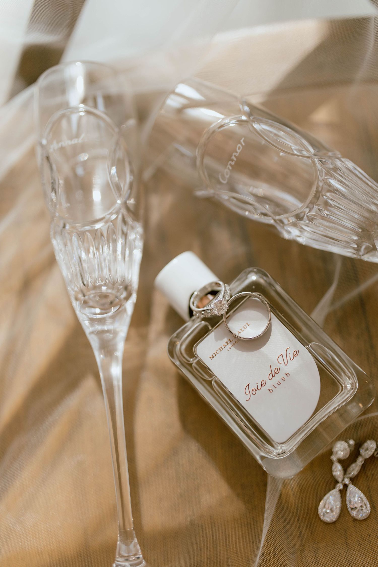 Wedding details with champagne glasses, perfume bottle and jewelry on a beige surface.
