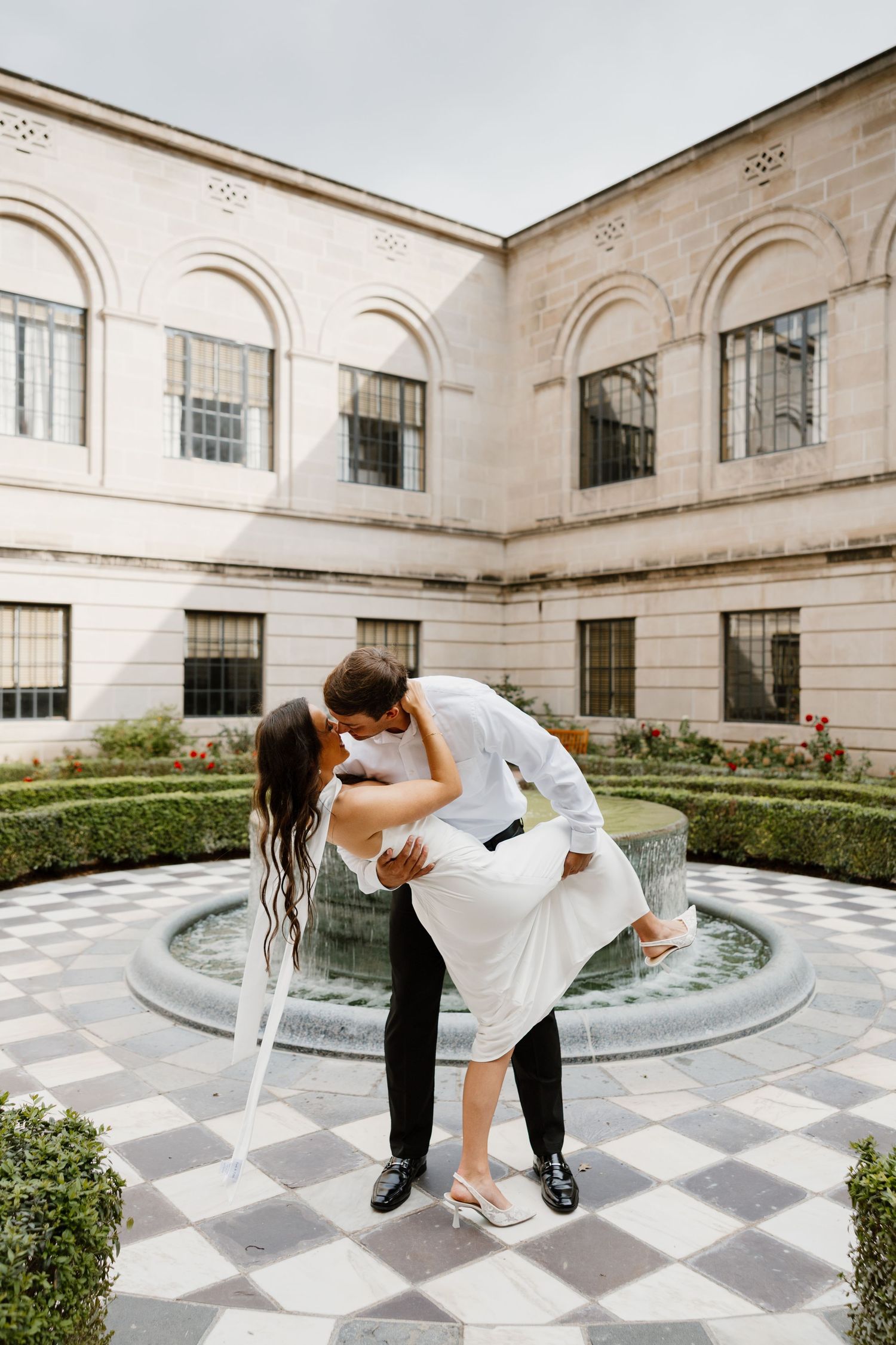 Dancing couple performs dramatic dip pose by ornate fountain in formal garden.