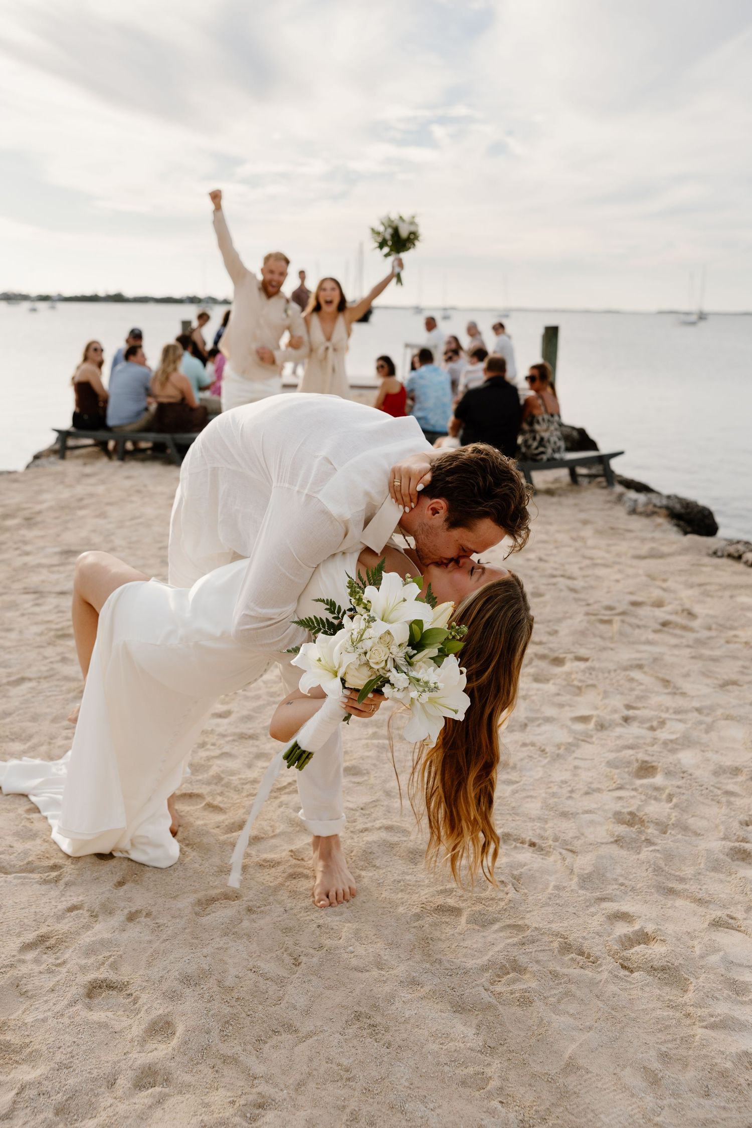 Groom dips bride for a romantic kiss on a sandy beach during their outdoor wedding ceremony by the water in Key Largo, Florida.