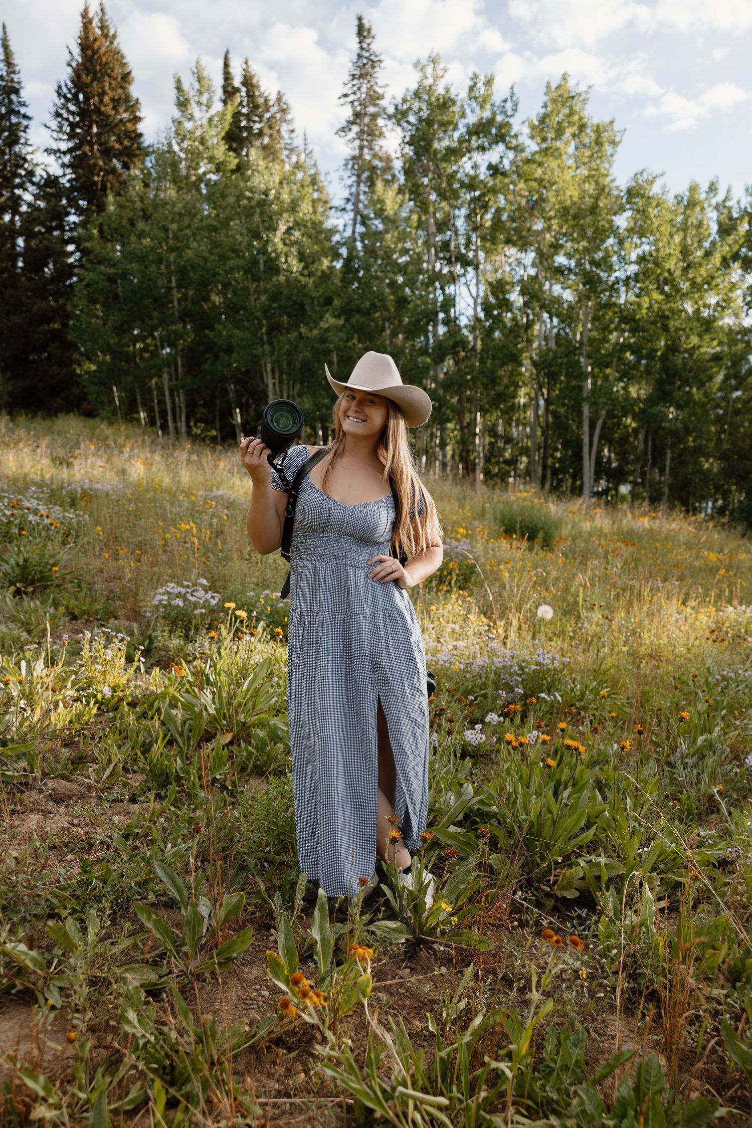 A photographer in a gingham dress and cowboy hat stands in a mountain meadow with aspen trees.
