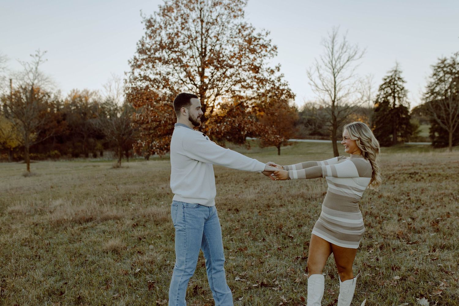 Two people hold hands and dance together in an autumn field during golden hour with trees in the background.