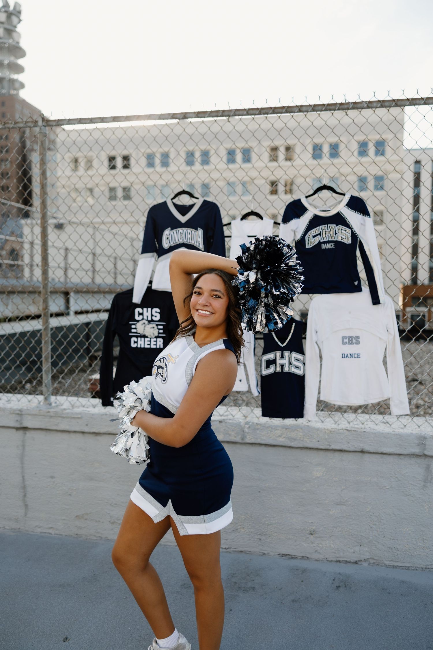 A cheerleader poses with pom-poms in front of a wall displaying navy and white sports uniforms and apparel.