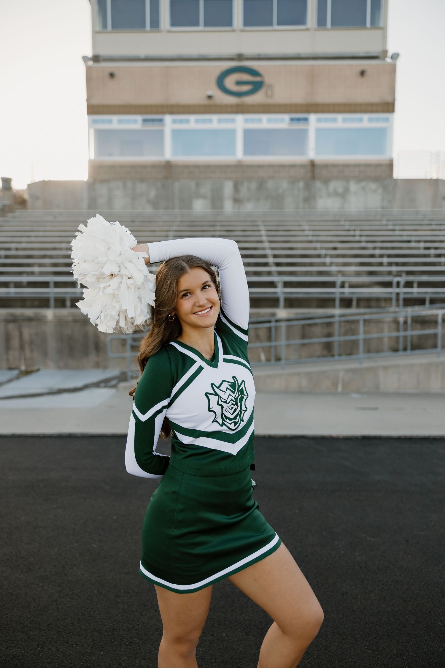 A cheerleader in green and white uniform with pom pom poses in front of empty stadium bleachers.