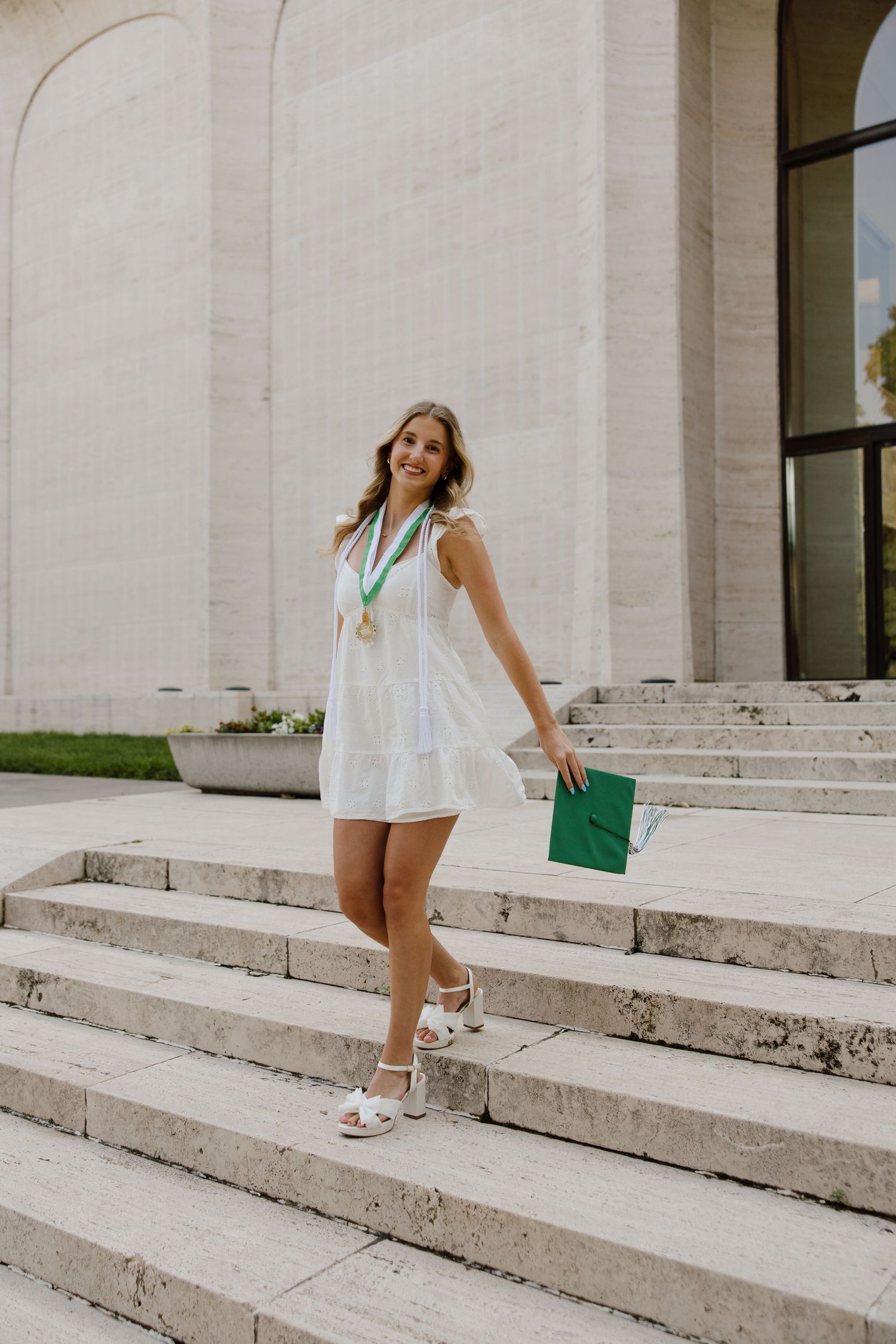 Graduate in white dress walks down concrete steps at graduation photoshoot.