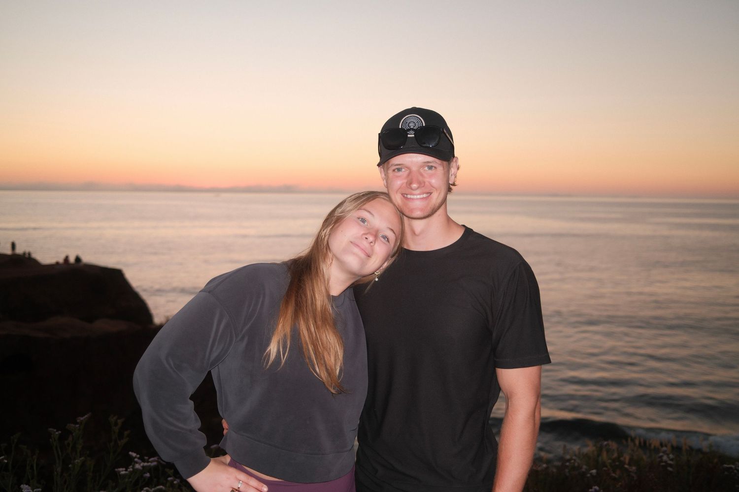 Couple smiling together at a scenic ocean overlook during a colorful sunset with warm pink and orange skies.