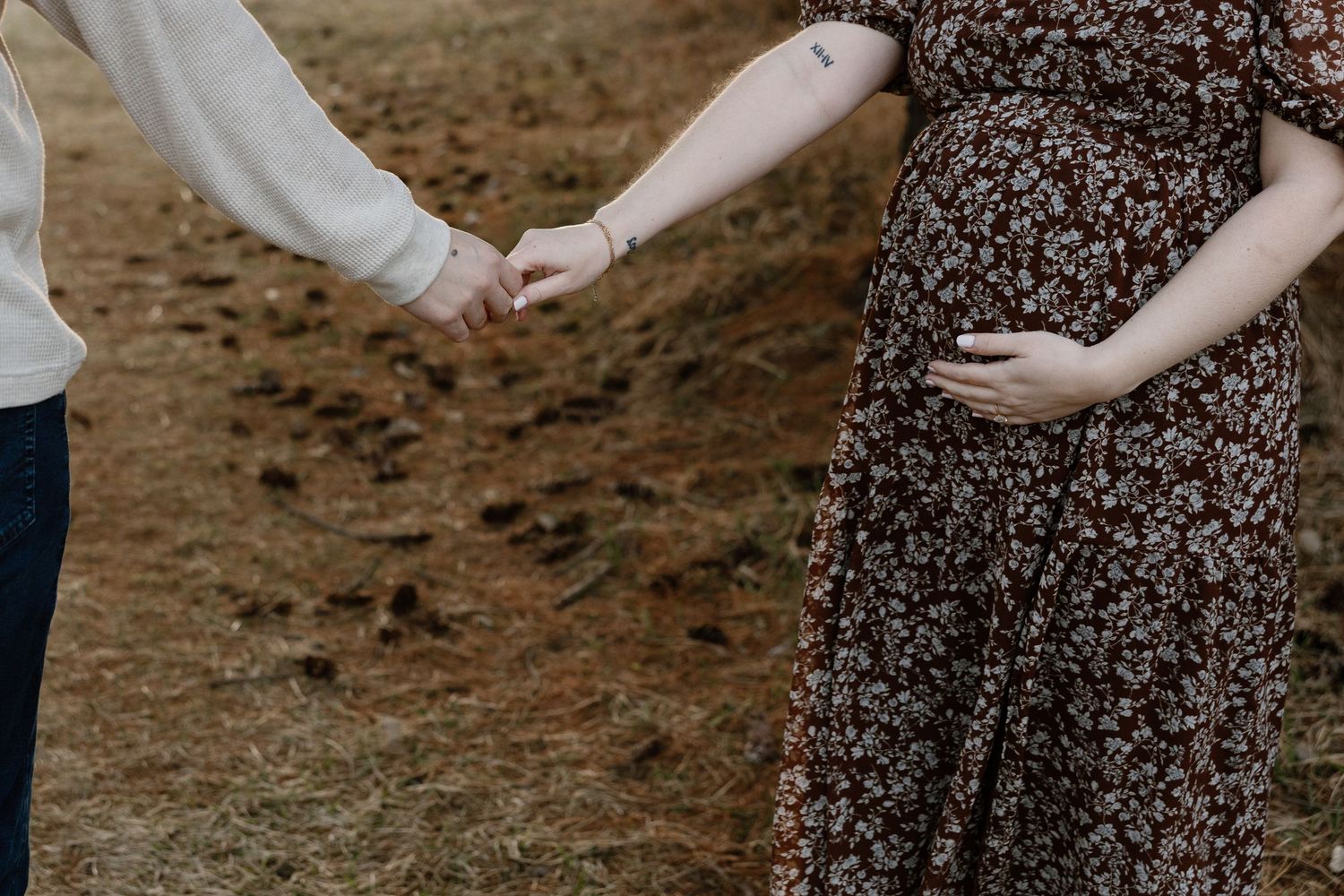 Pregnant woman in floral dress holding hands with partner outdoors on brown grass field.
