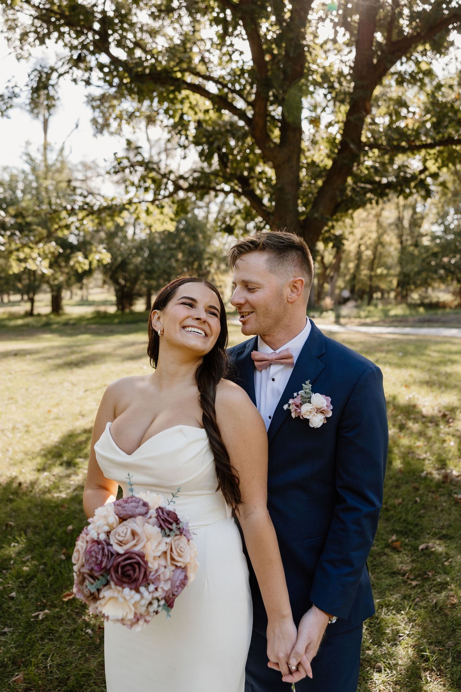 Joyful wedding couple laughing together in sunlit garden setting with pink bouquet.