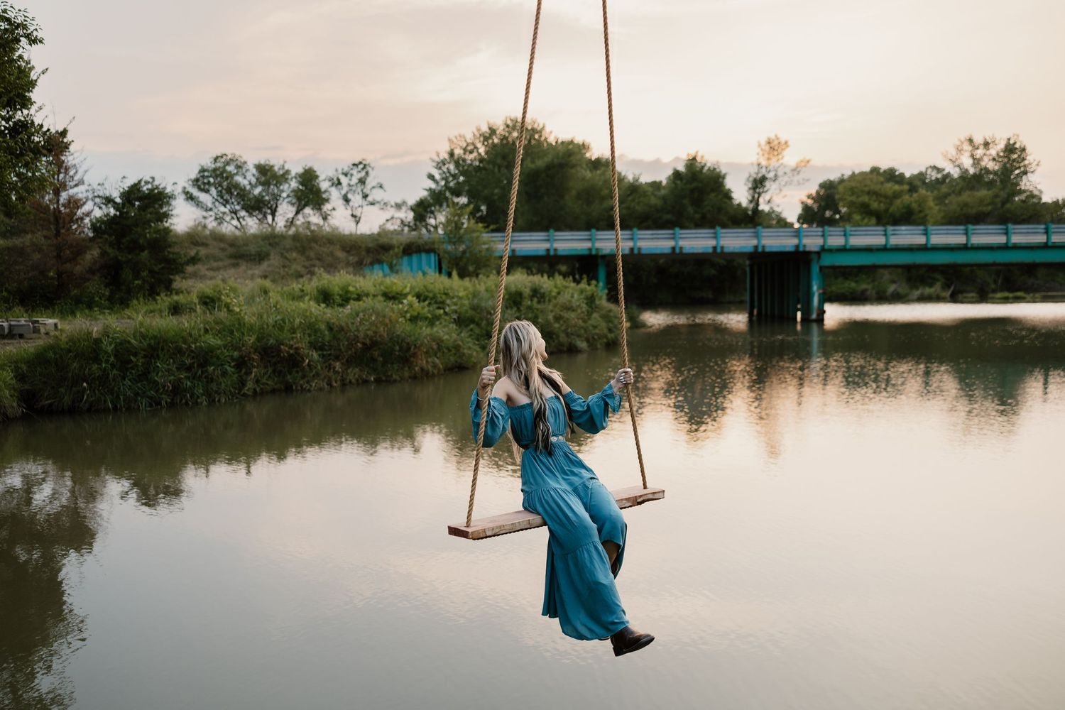 Person in blue dress swinging over a tranquil river at sunset.
