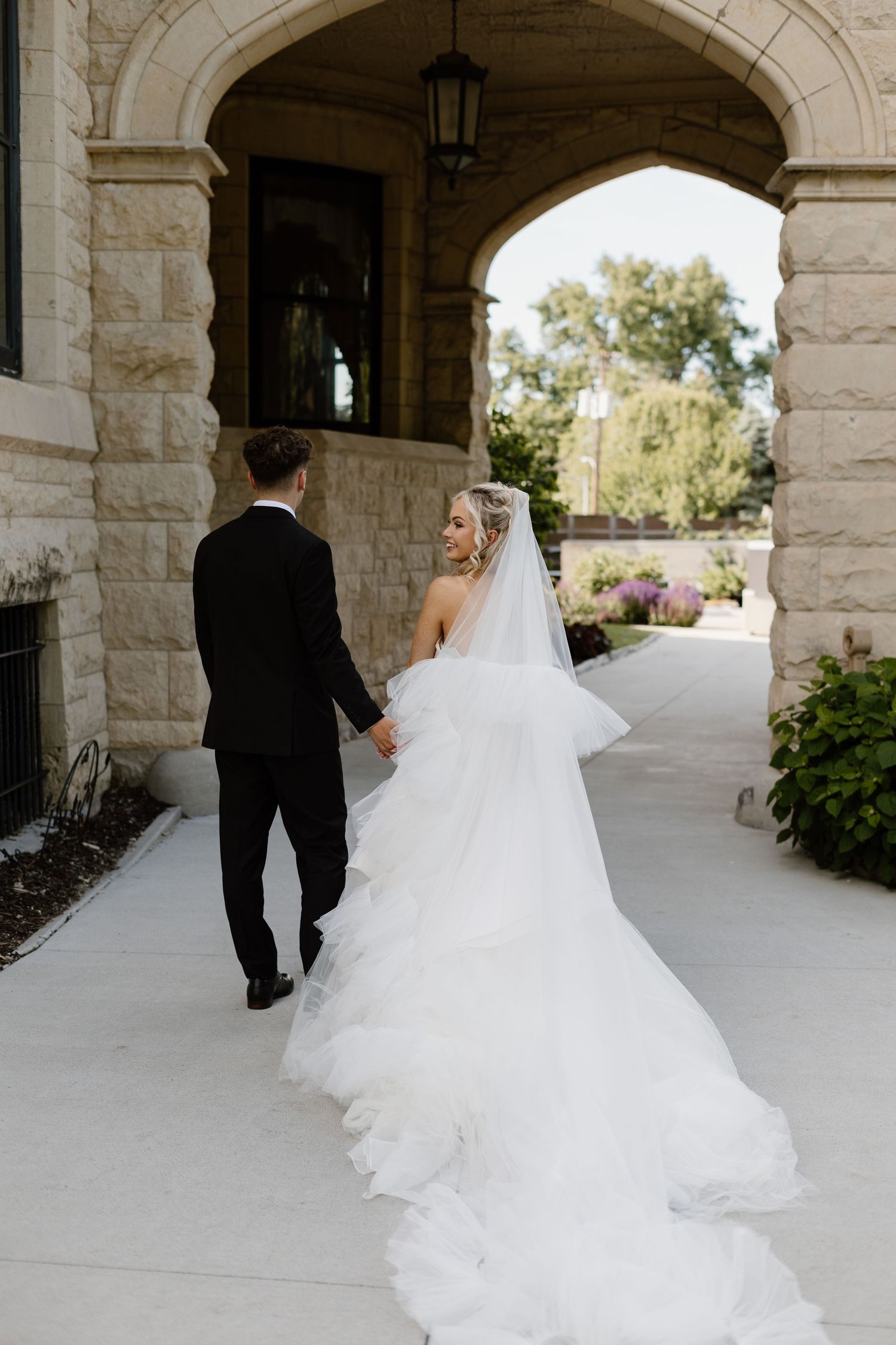 Bride in flowing white gown and veil walks hand-in-hand with groom in black tuxedo under stone arch.