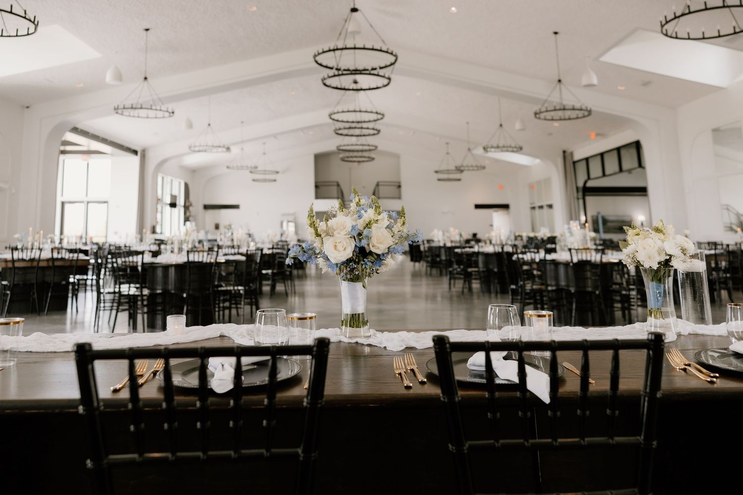 Elegant wedding reception venue with black chairs and white floral centerpieces in a bright modern banquet hall.