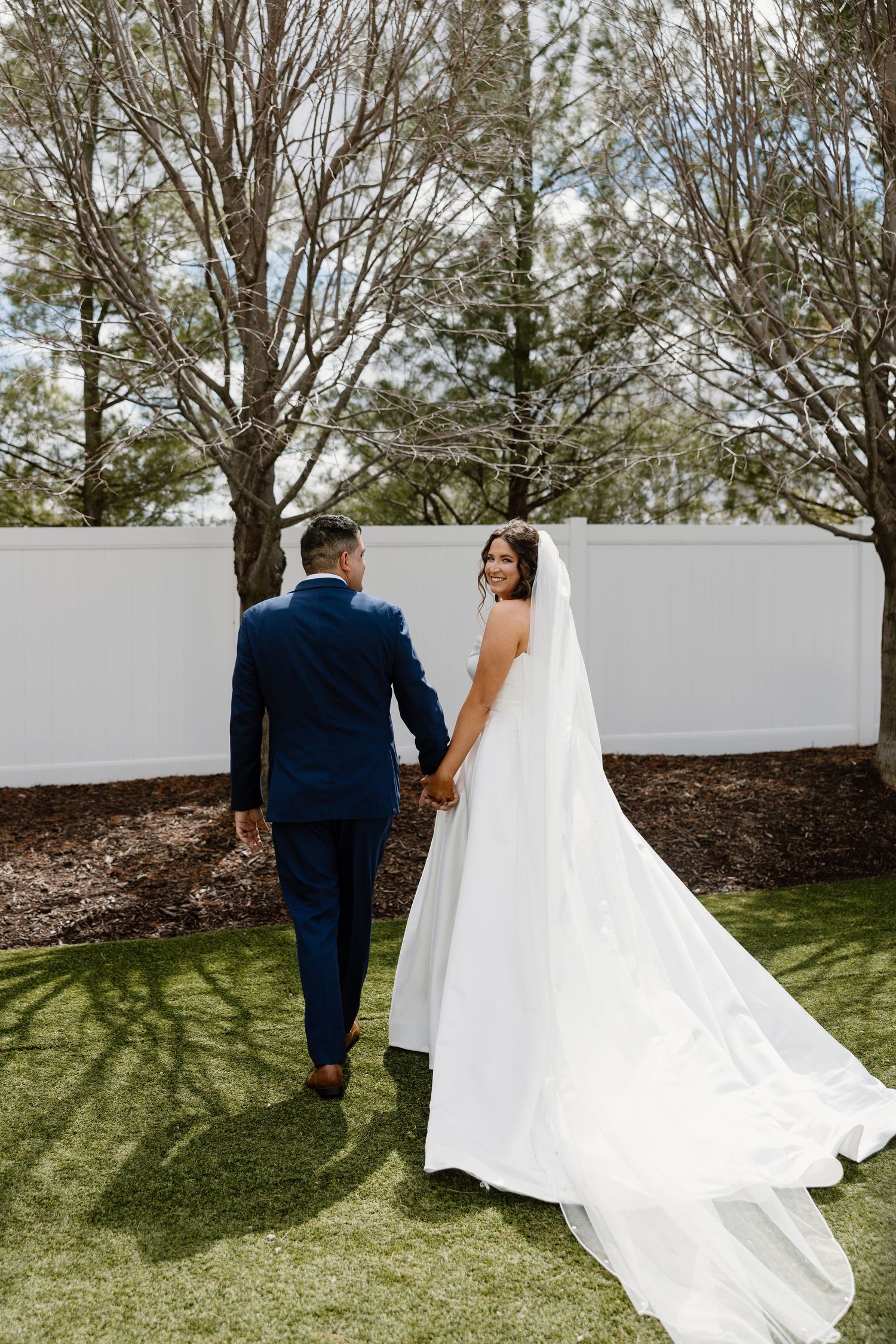 A bride and groom walk hand in hand through a garden, the bride wearing a flowing white gown with a long veil.