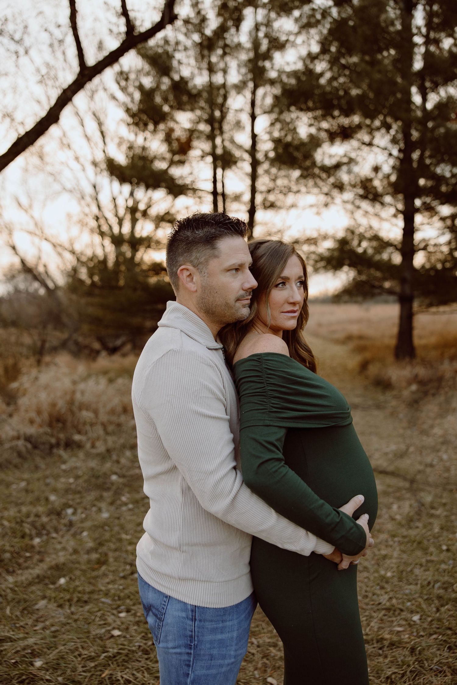 Romantic maternity portrait of couple embracing among pine trees at dusk.