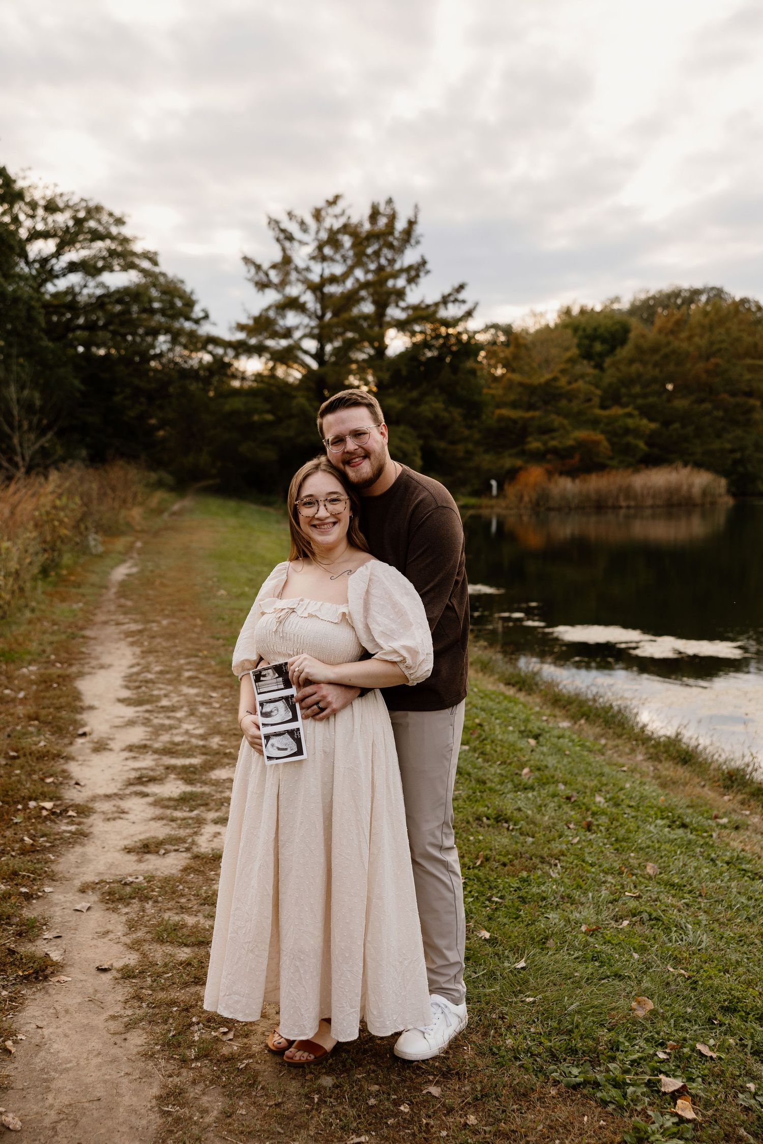 Expecting couple sharing tender moment by lakeside while displaying ultrasound images.