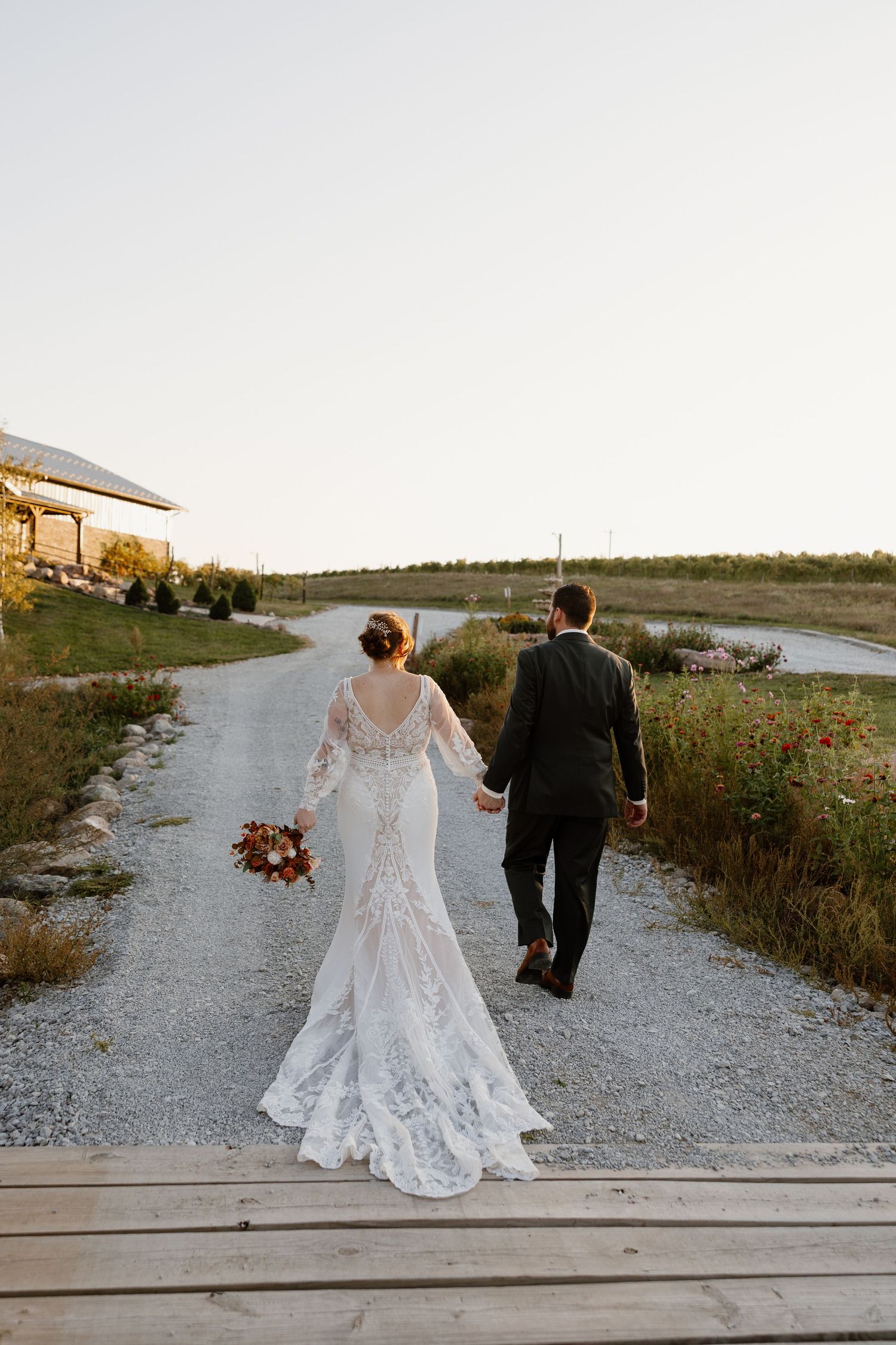 Couple walking along boardwalk at sunset, bride in lace dress holding orange and white bouquet.
