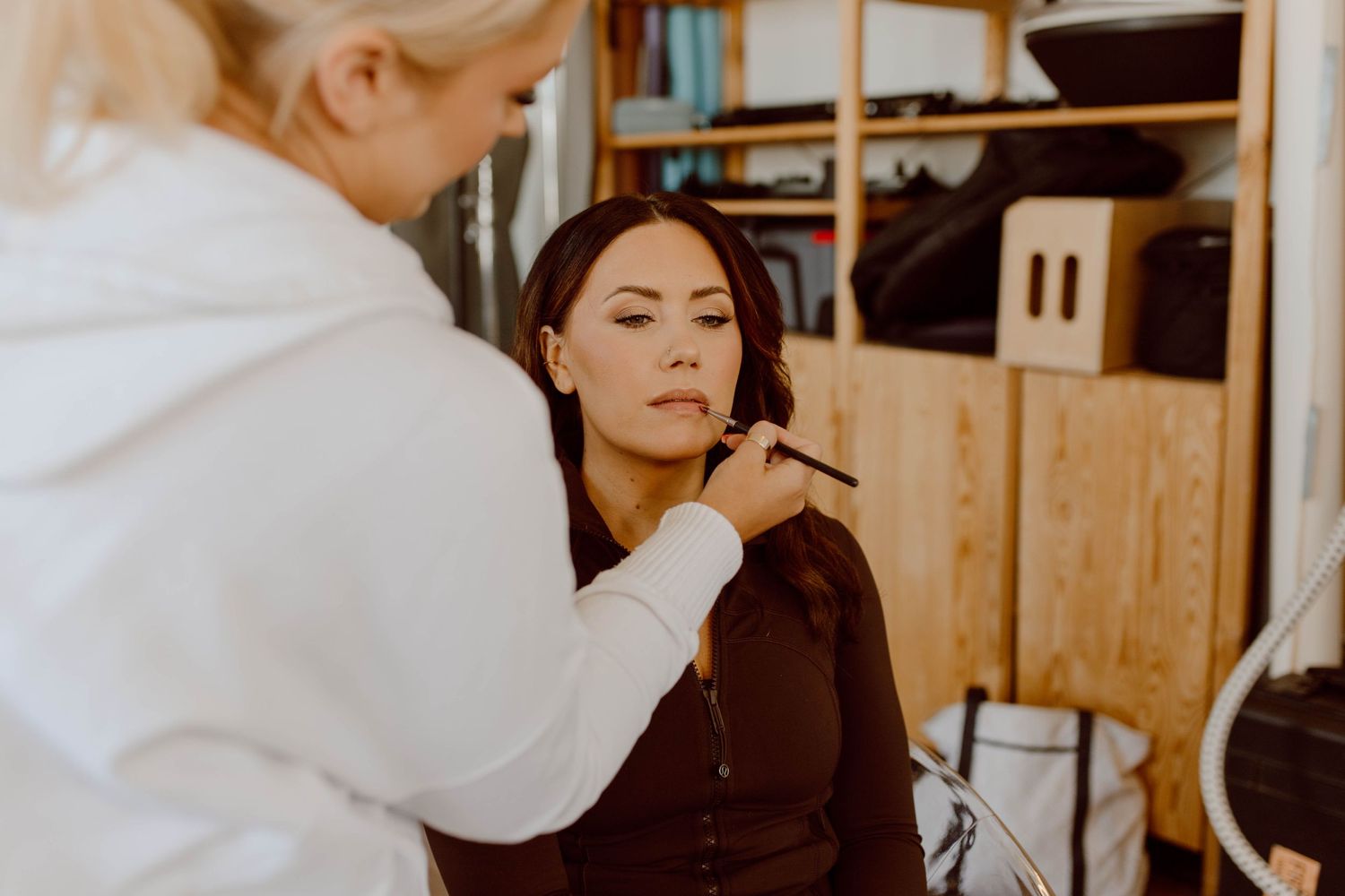 Makeup artist in white sweater applies cosmetics to client in warm-toned studio setting.