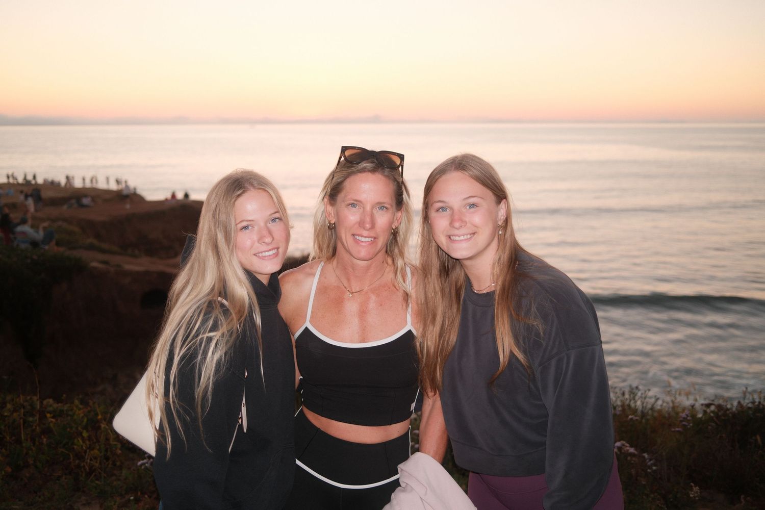 Three smiling women pose together at a scenic ocean overlook during a beautiful sunset.