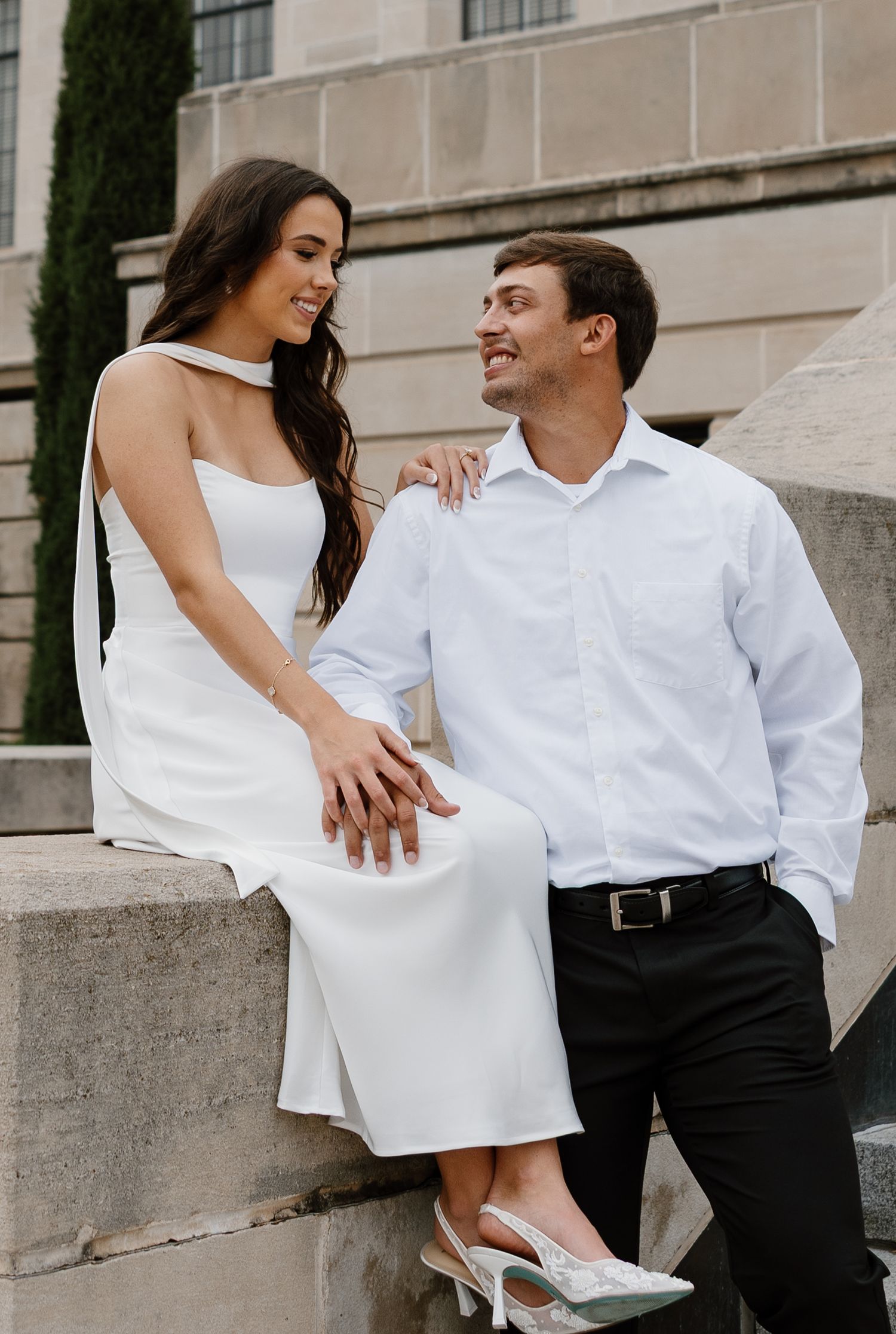 A couple in formal attire poses playfully on architectural steps with white stone details.