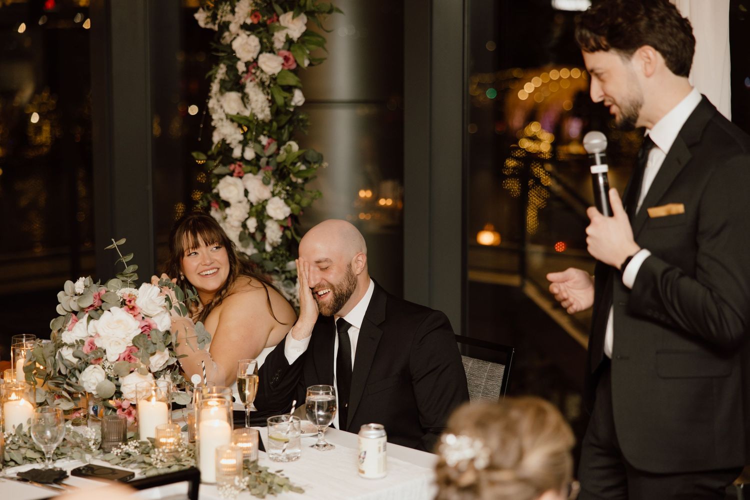 Reception dinner guests laugh together at wedding speeches while seated at elegantly decorated table.