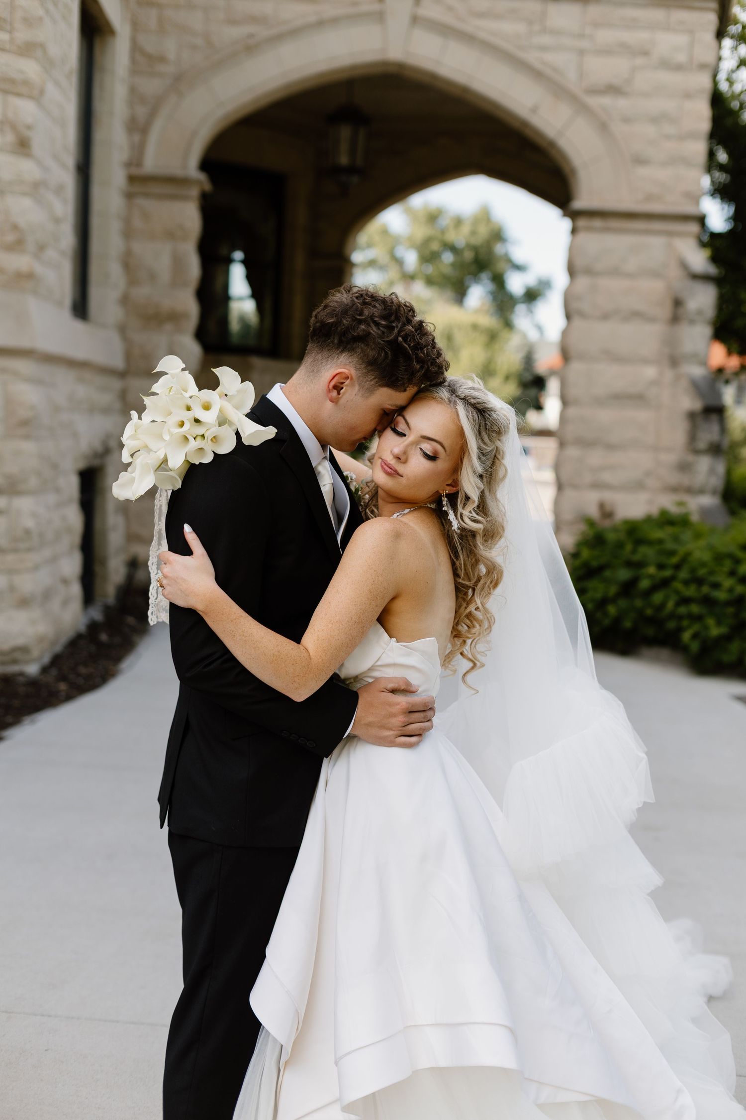 A  Nebraska bride and good embrace tenderly outside Joslyn Castle, she holds white calla lilies in a flowing wedding gown.