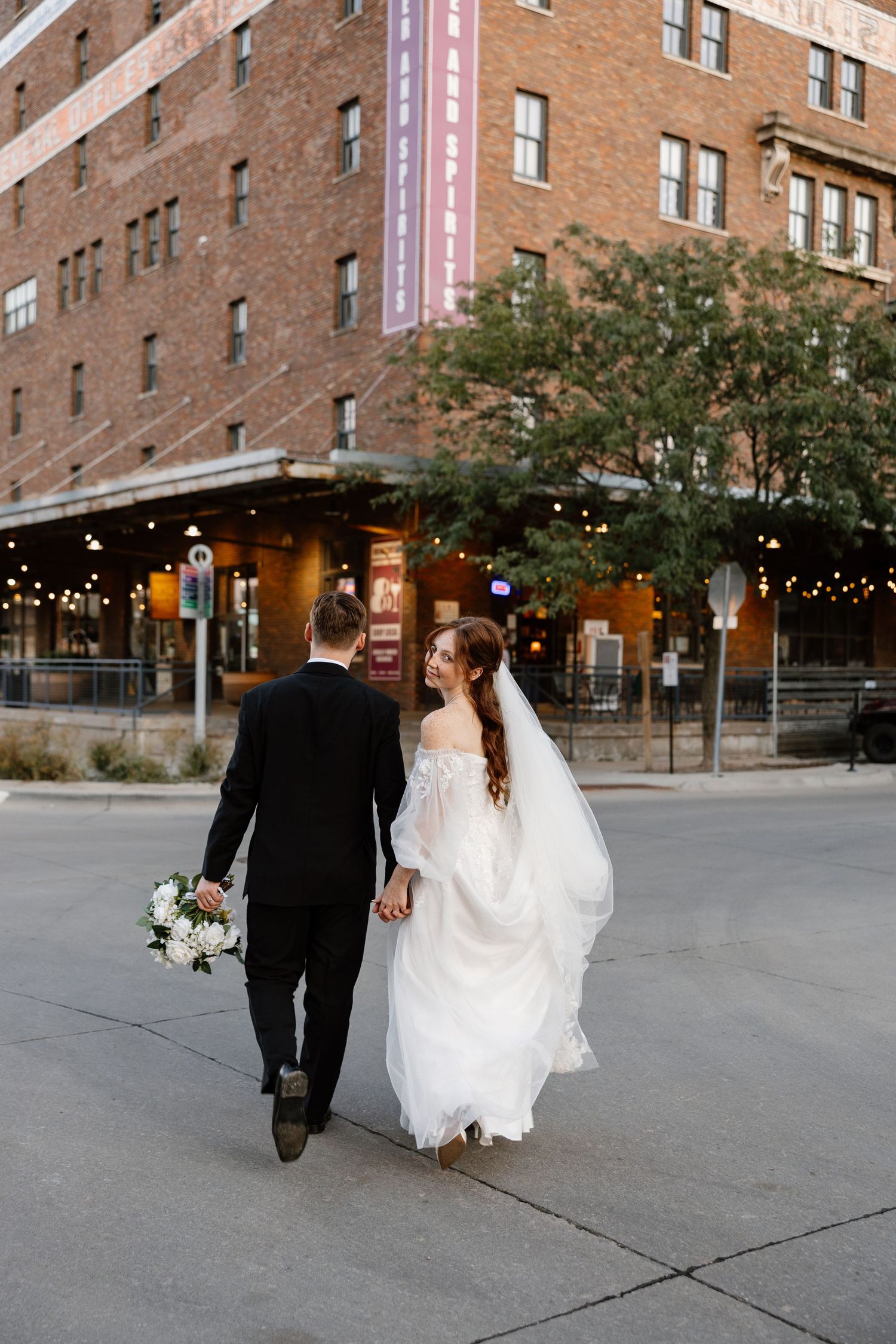 Newlyweds walking together along urban brick building with string lights in downtown omaha