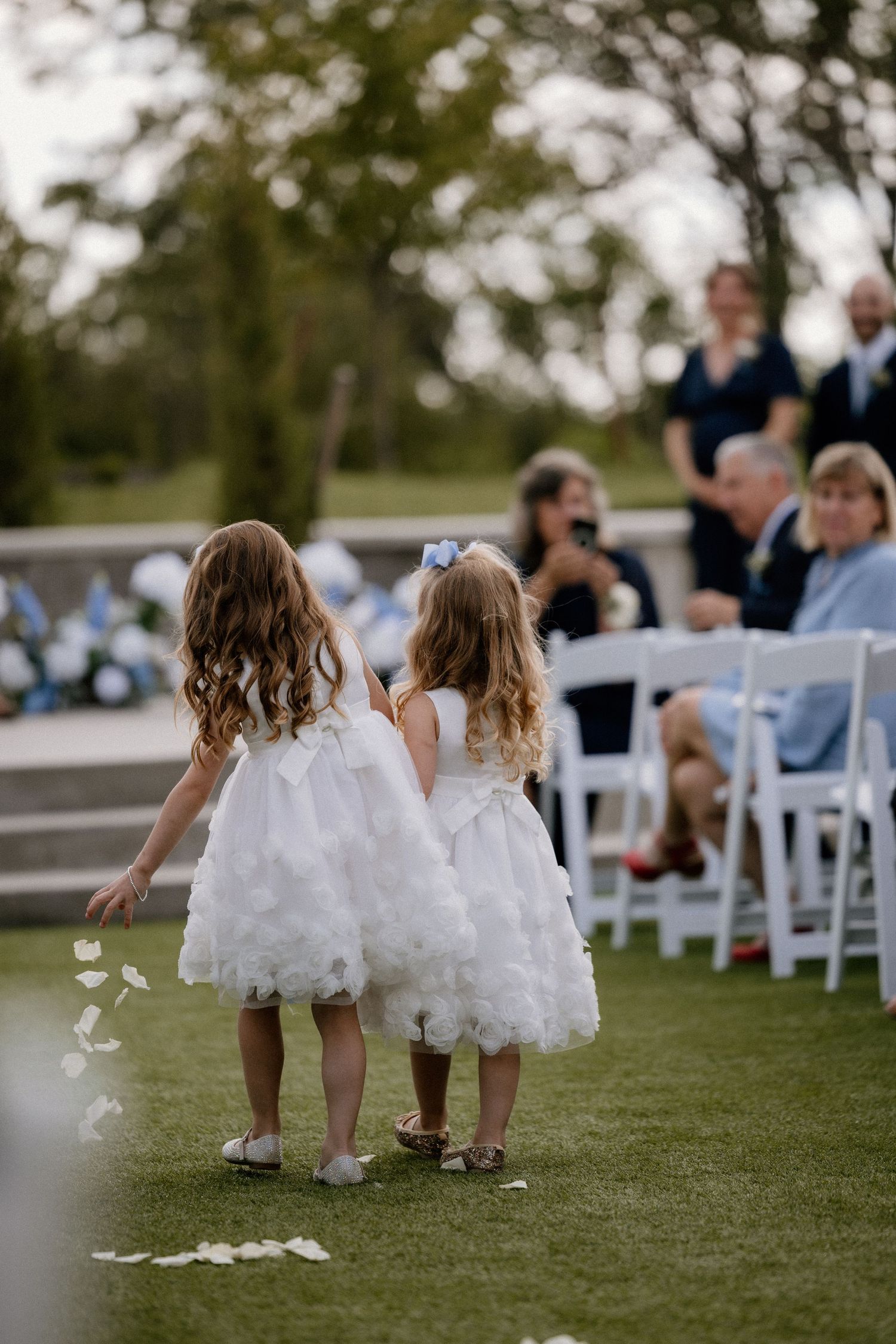 Two flower girls in white dresses scatter petals down the aisle at an outdoor wedding ceremony.