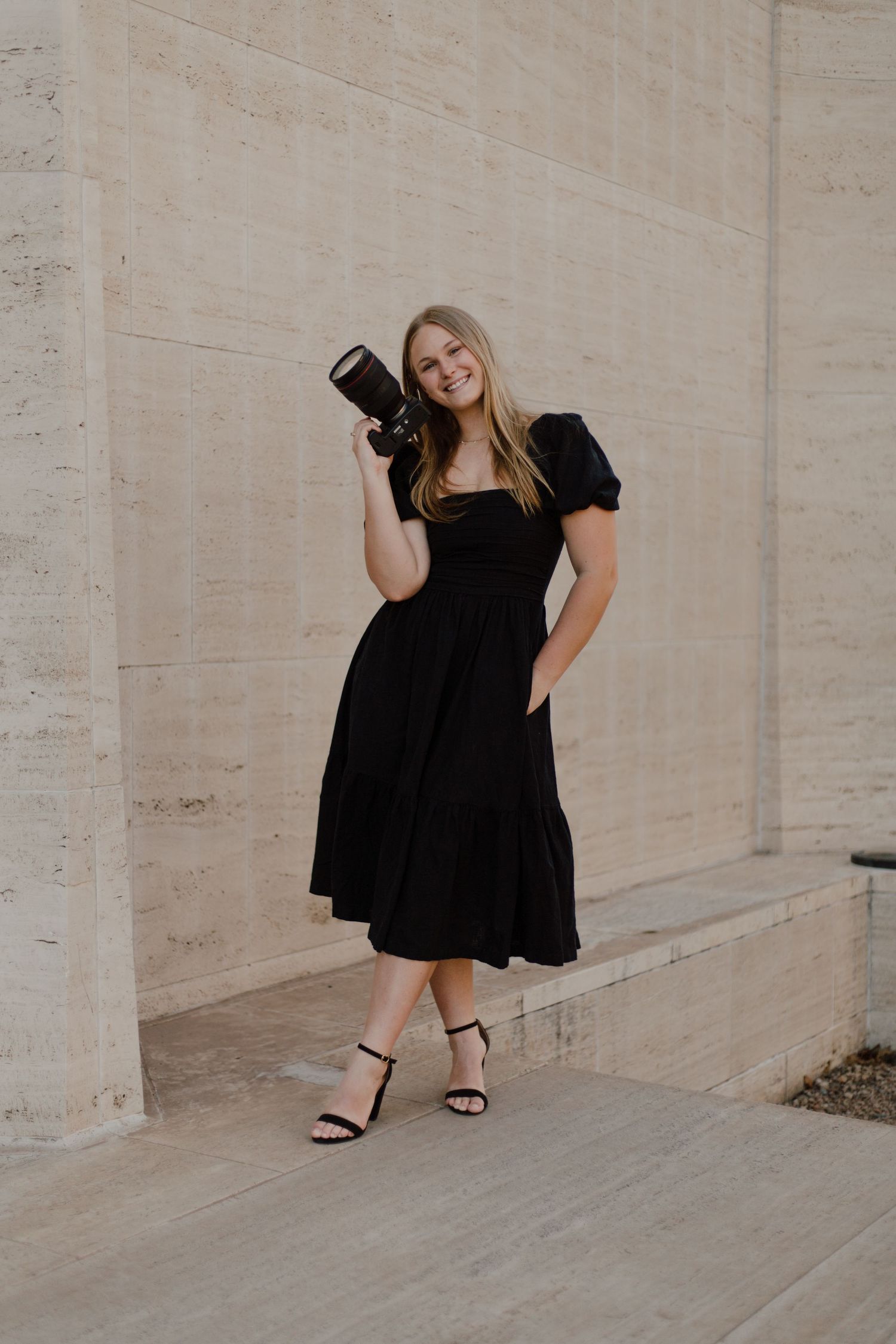 A photographer in a black midi dress and heels poses with a professional camera against a concrete wall.