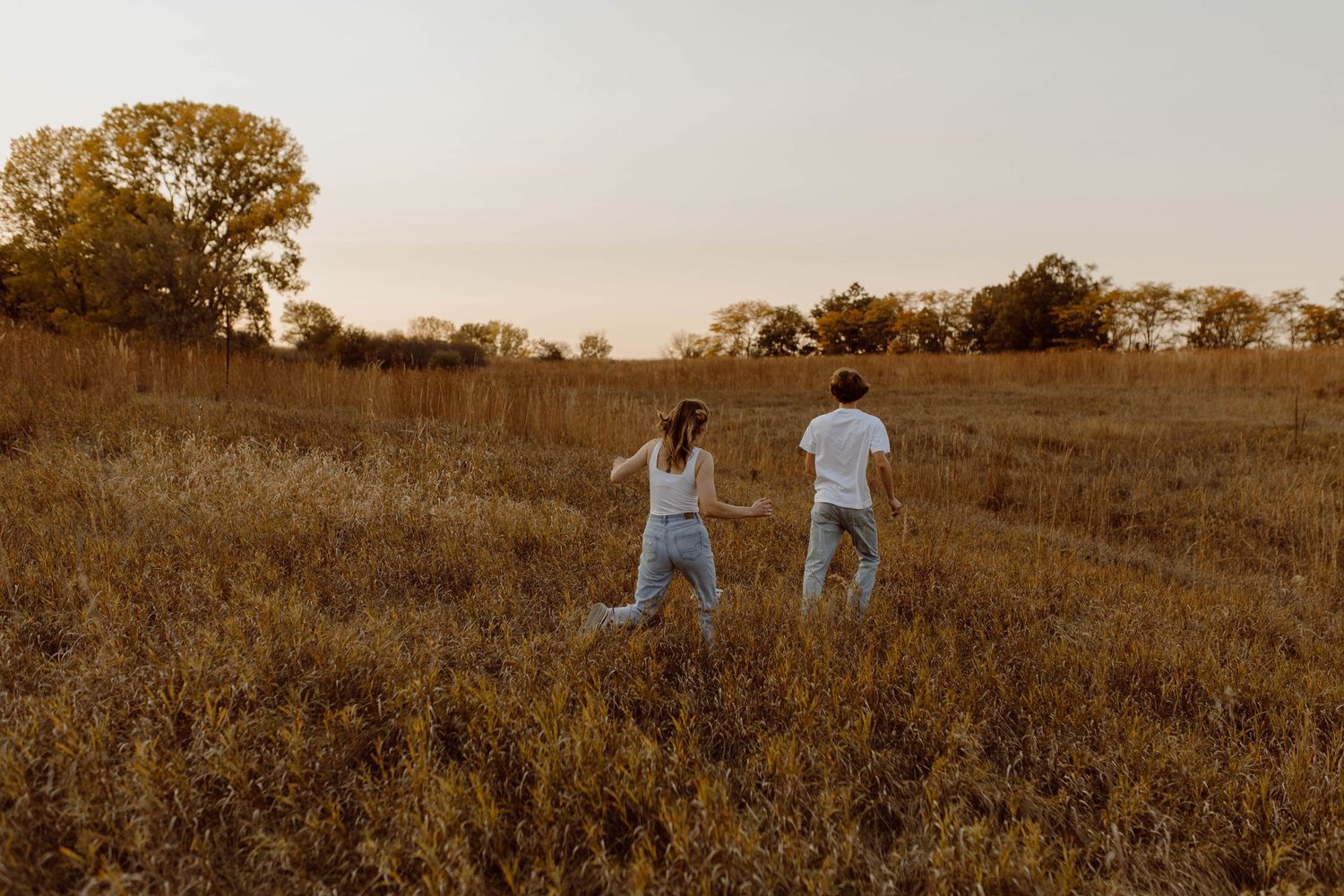 Two people walking together through a golden sunlit meadow at sunset with trees in the background.