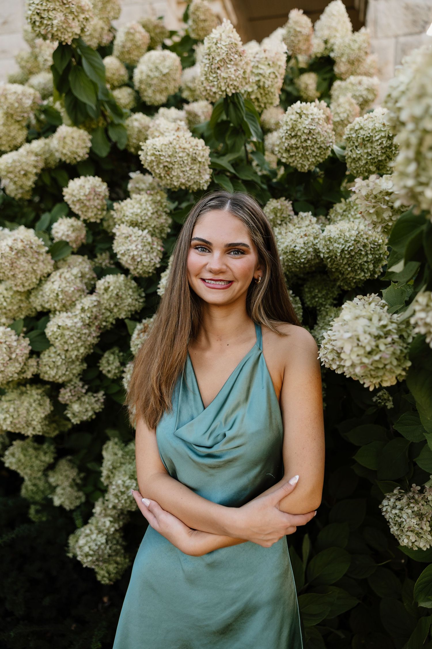 A woman in a sage green dress poses against a backdrop of white hydrangea flowers.