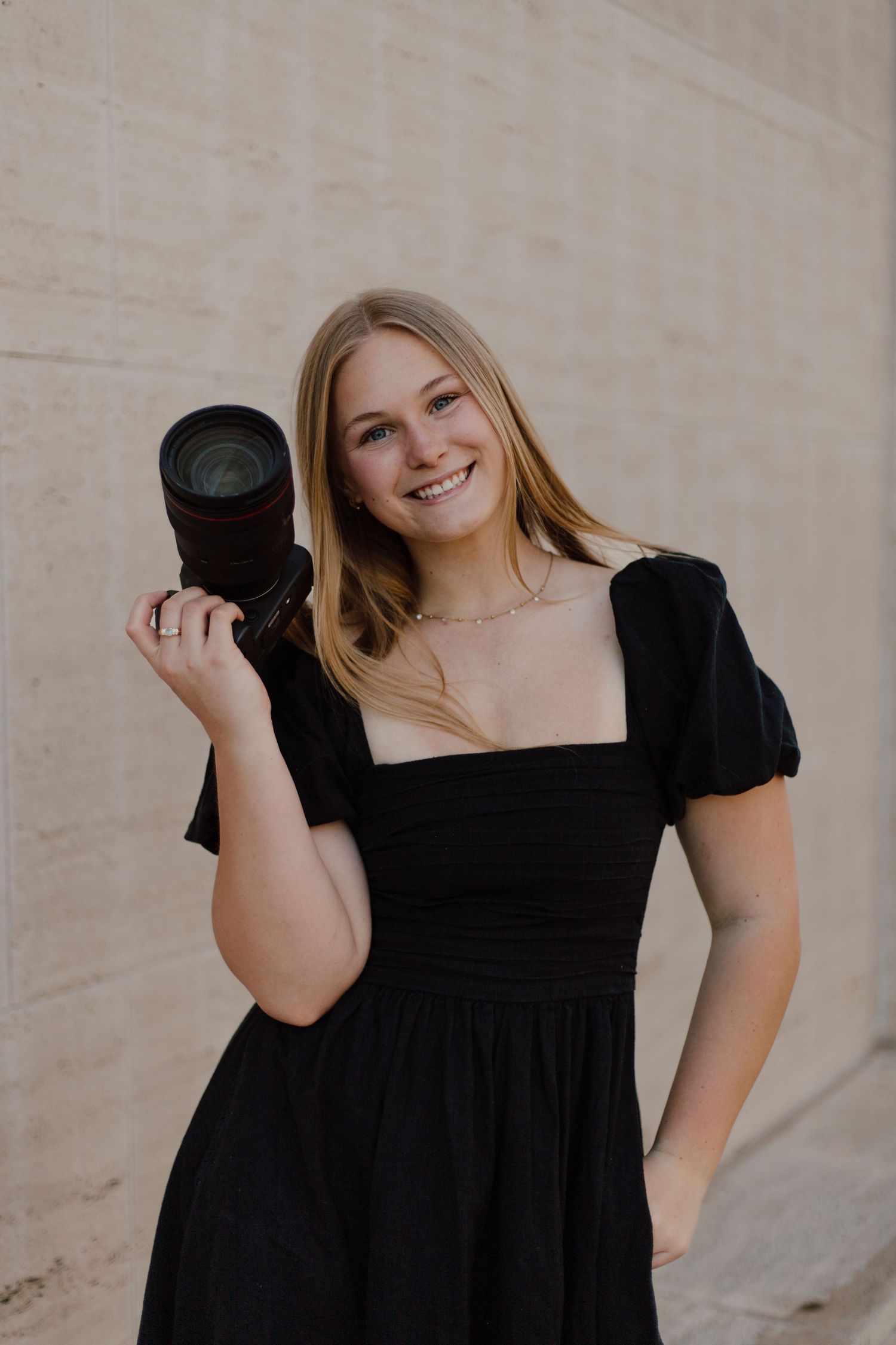 A photographer in a black dress holds up a professional camera lens while posing against a beige wall.