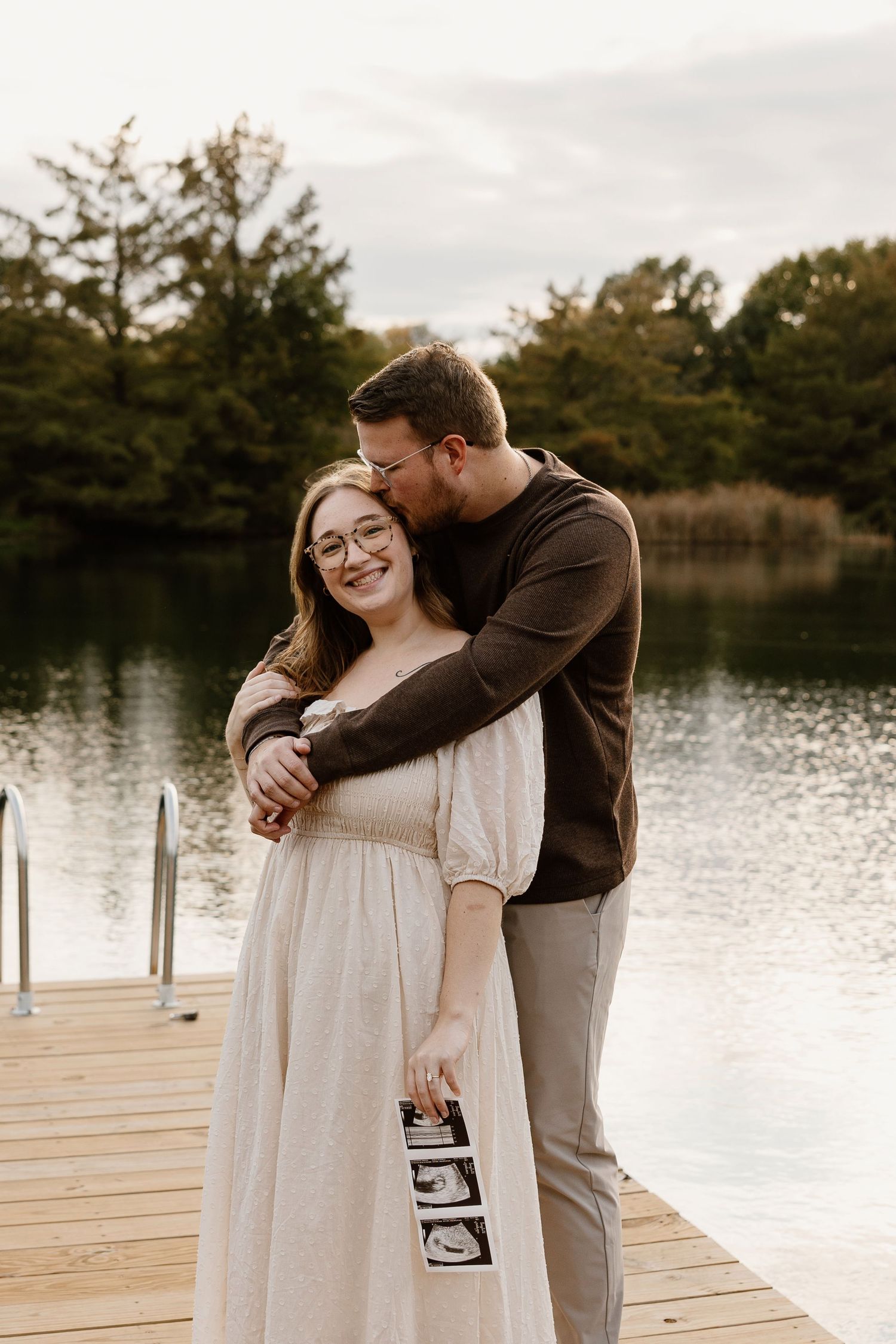 Couple embracing on wooden dock by lake while holding ultrasound photos.