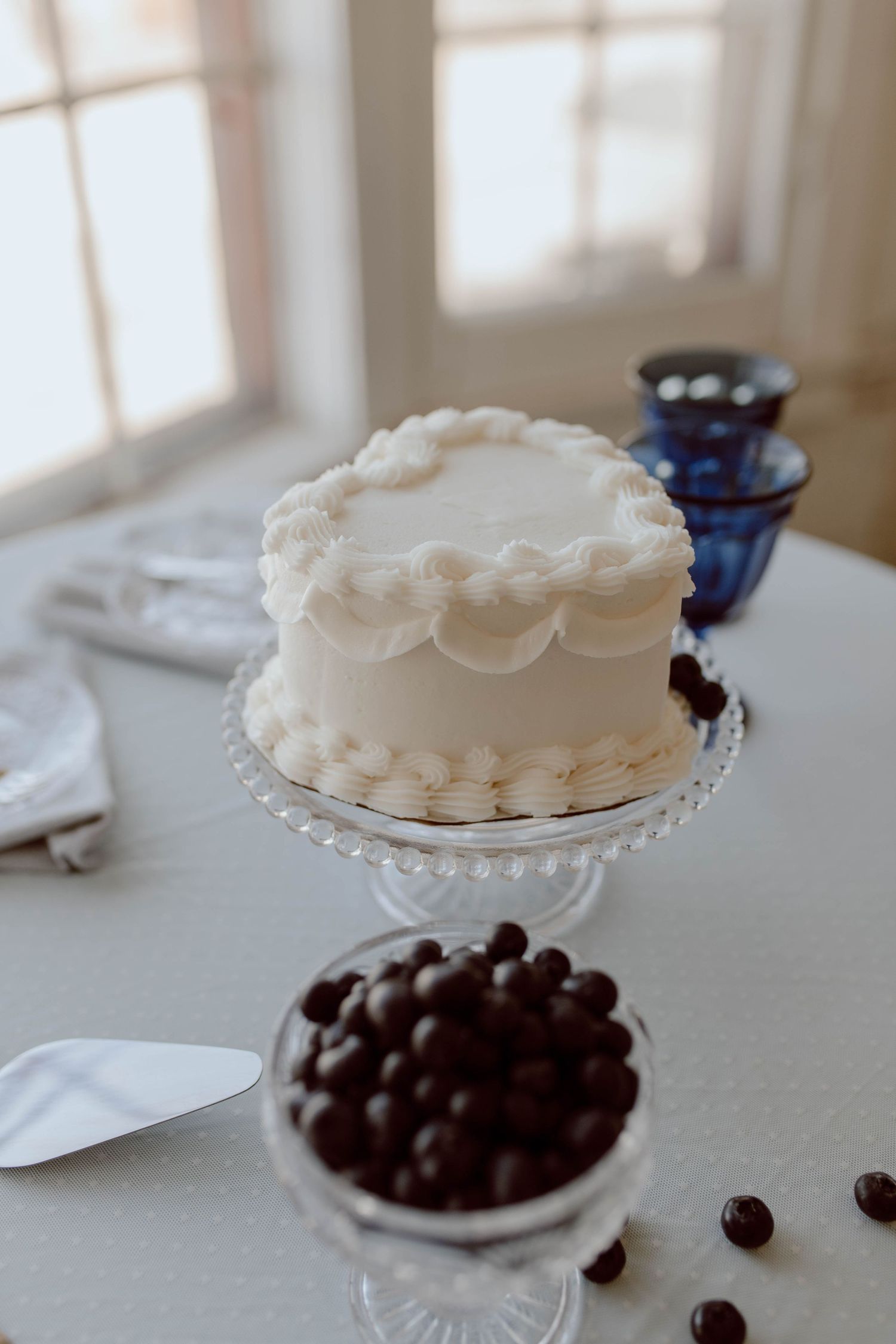 A white frosted cake with scalloped decorations sits on a crystal cake stand beside a bowl of chocolate truffles.