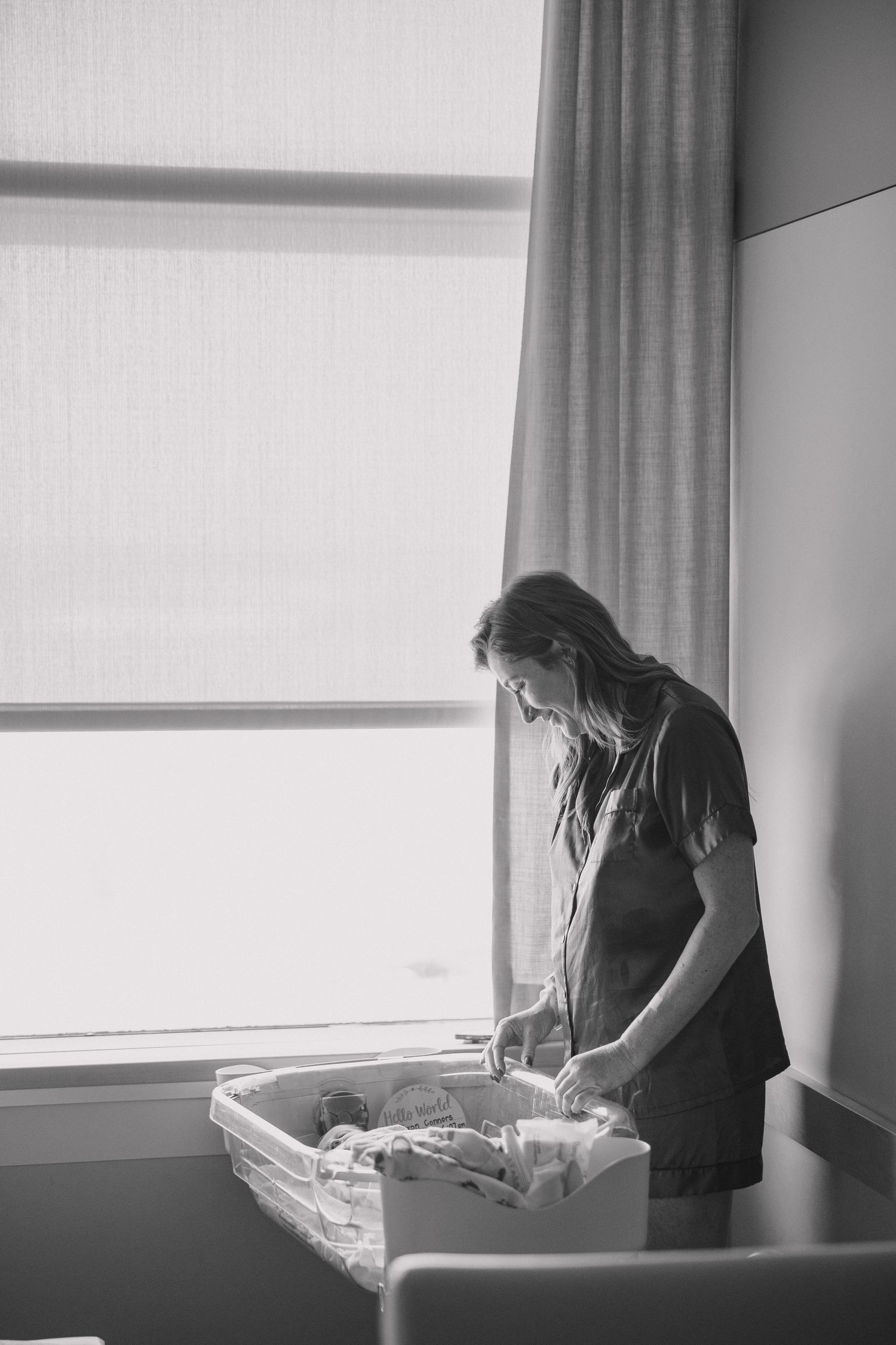 Woman standing by window in dark room tending to contents of a bassinet in black and white photography.