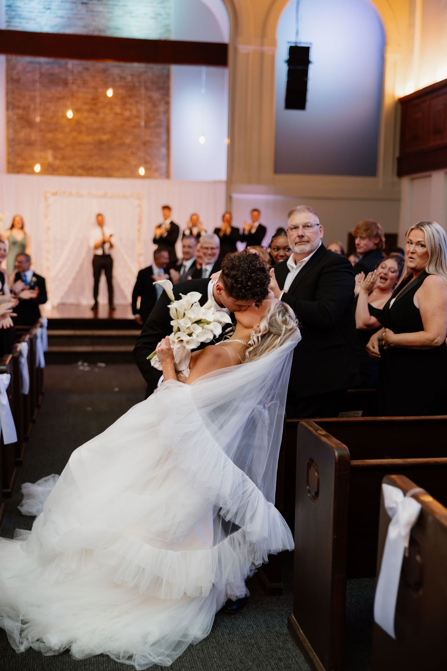 Bride and groom share a romantic dip kiss in church aisle while wedding guests cheer and applaud during ceremony.