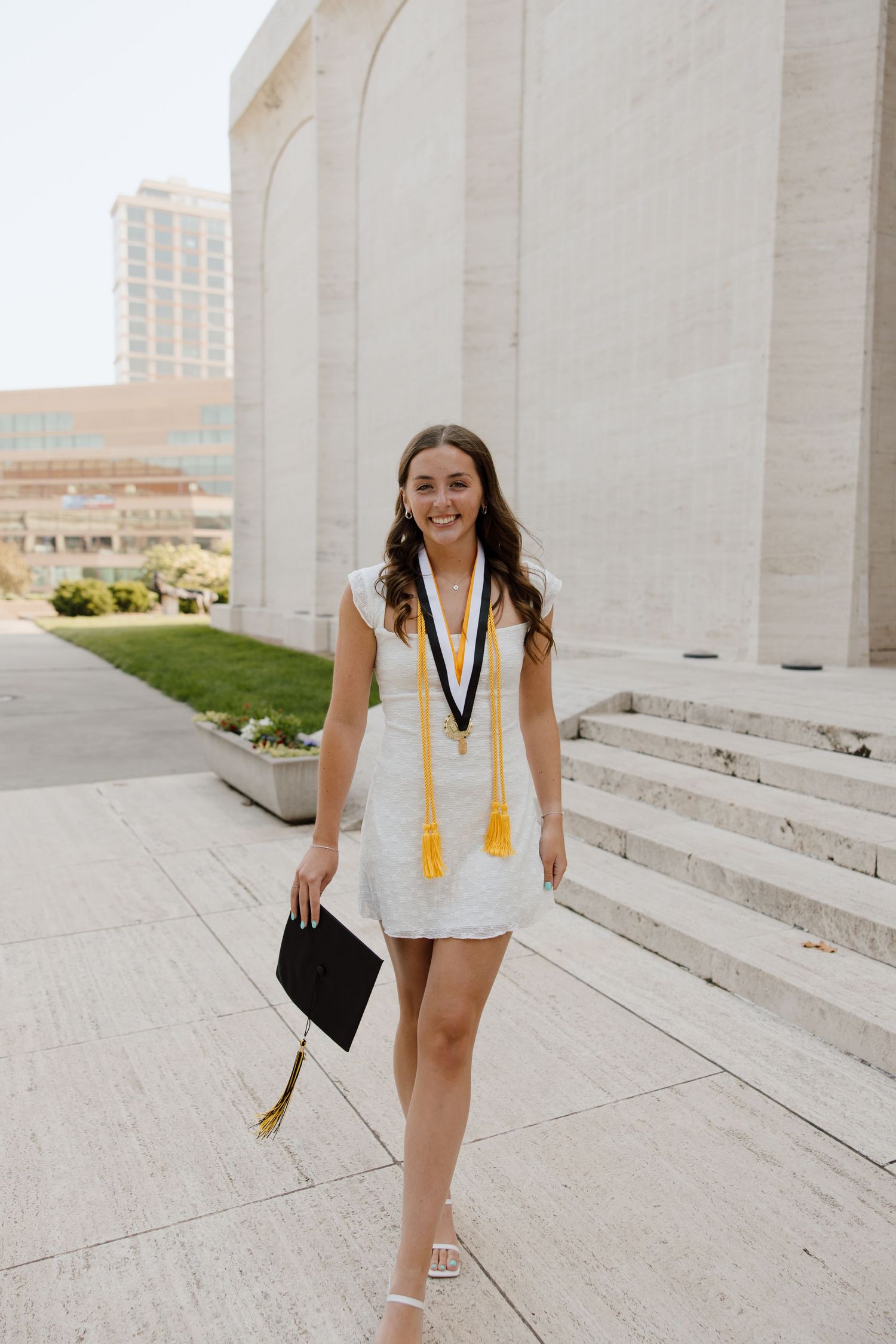 Graduate in white dress and academic stole poses on concrete steps of university building.