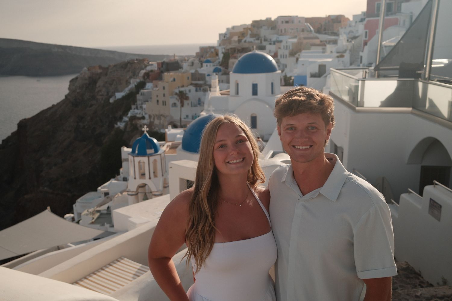 Couple smiling in Santorini Greece with iconic blue domed churches and white buildings overlooking the caldera at sunset.
