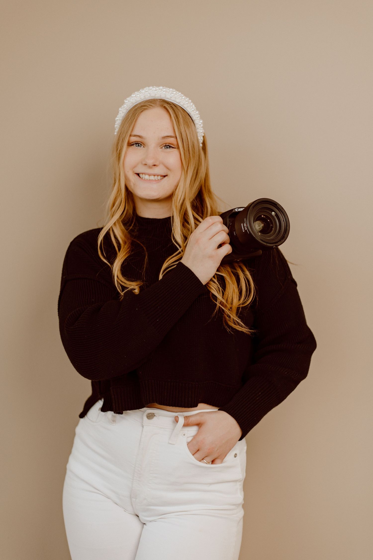 A photographer in a black sweater and white jeans holds a professional camera against a neutral backdrop.