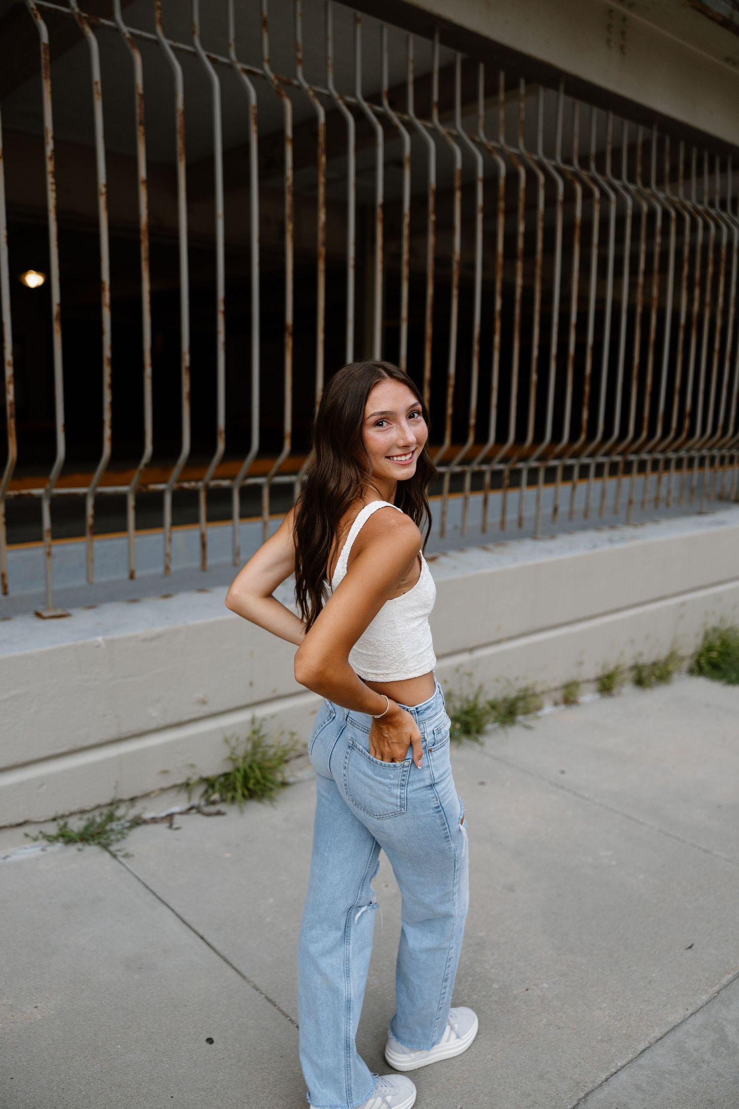 Person in light blue jeans and white crop top poses playfully against an urban railing background.