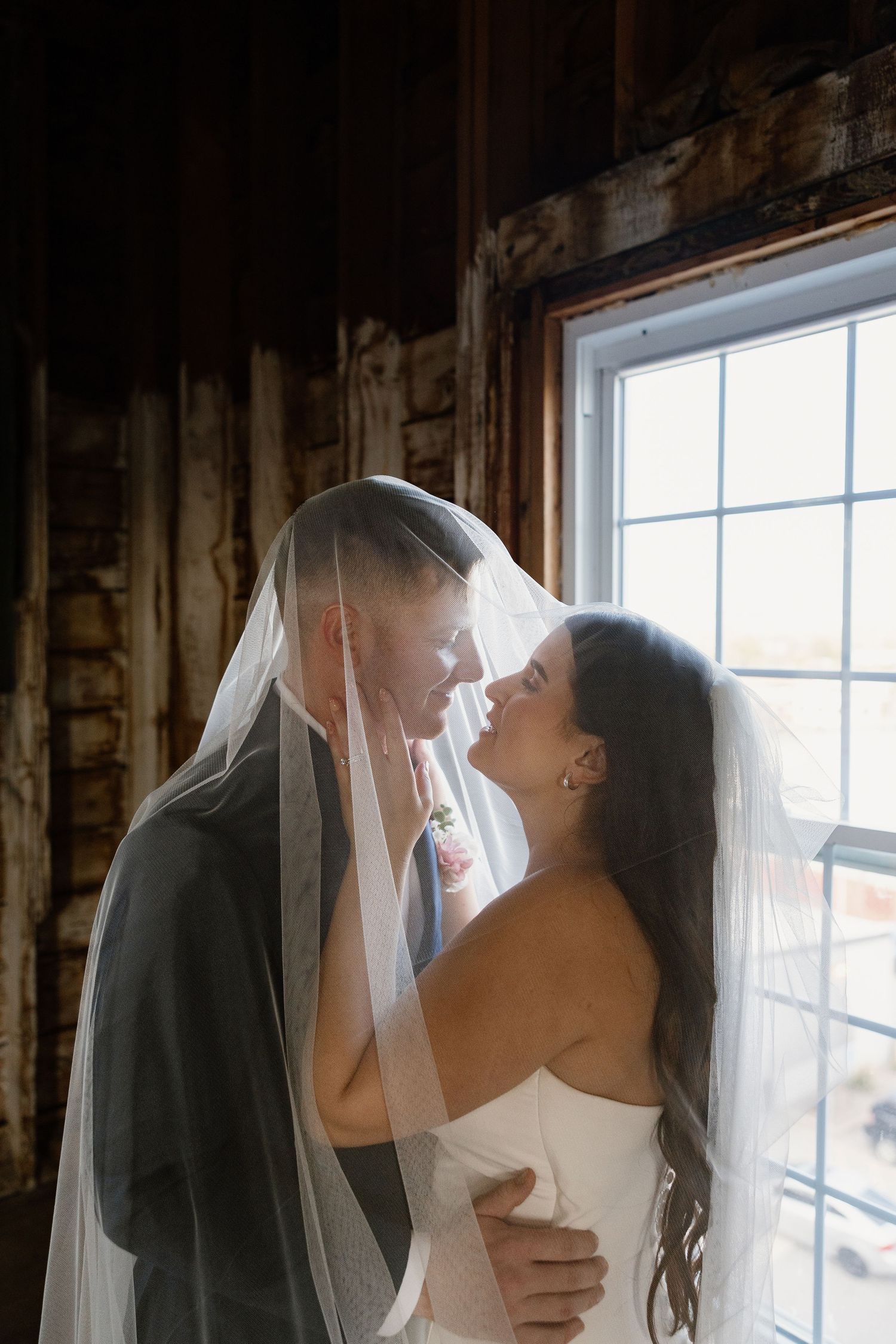 Intimate wedding moment captured by a window with soft natural light and flowing veil.