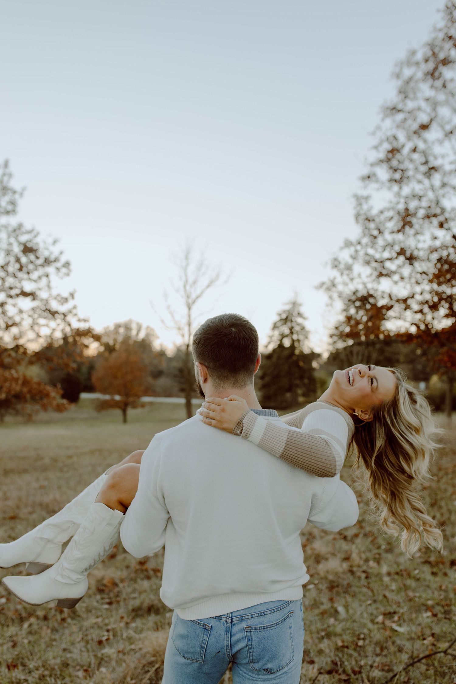 A couple shares a romantic moment in a park during autumn as he carries her while she leans back joyfully.