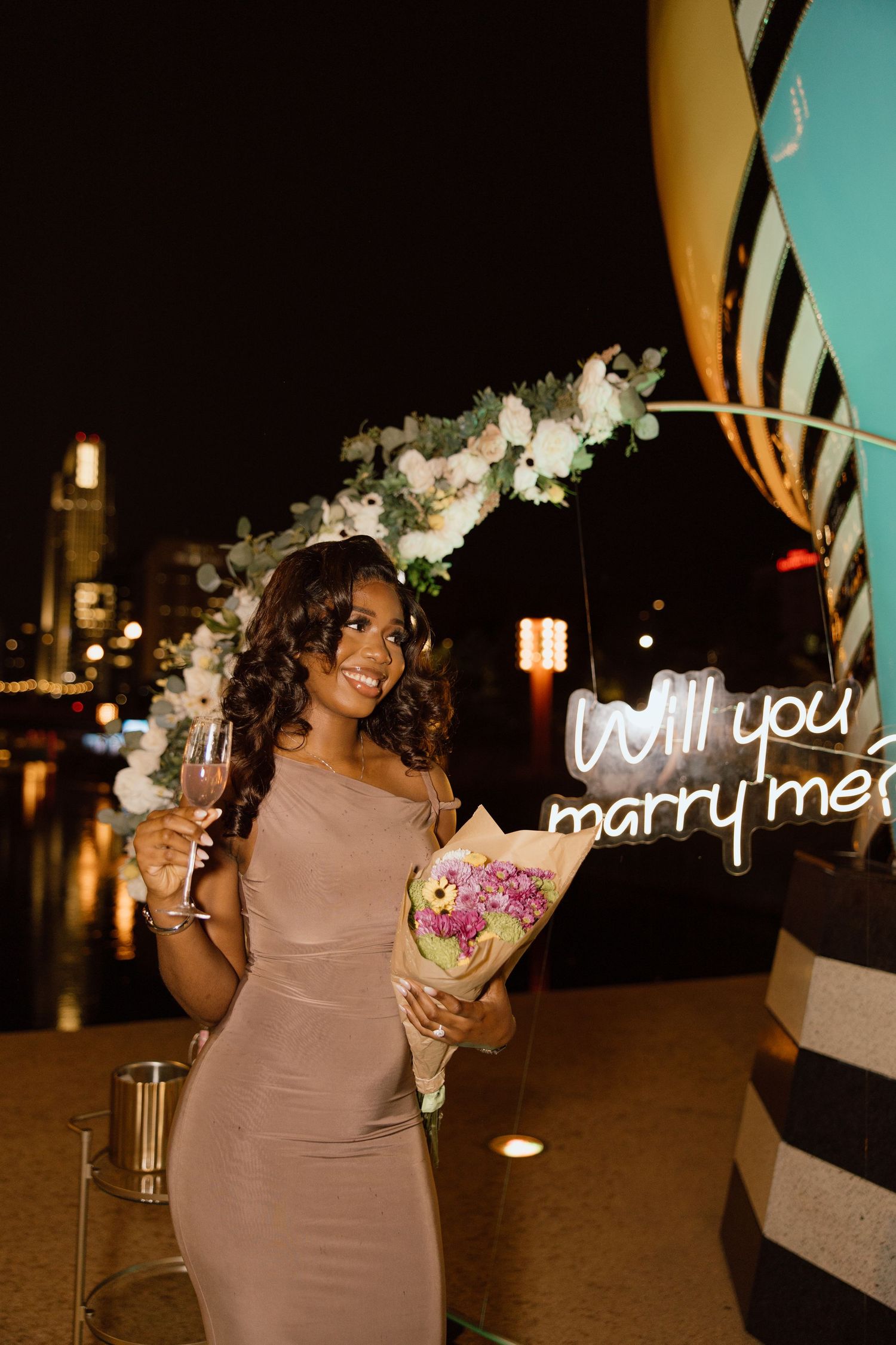 Woman in a mauve dress poses with illuminated 'Will you marry me?' sign at night.