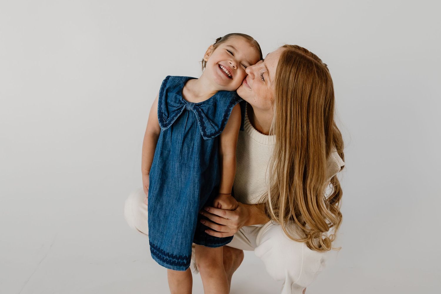 A woman in white kisses the cheek of a laughing little girl in a blue denim dress against a white background.