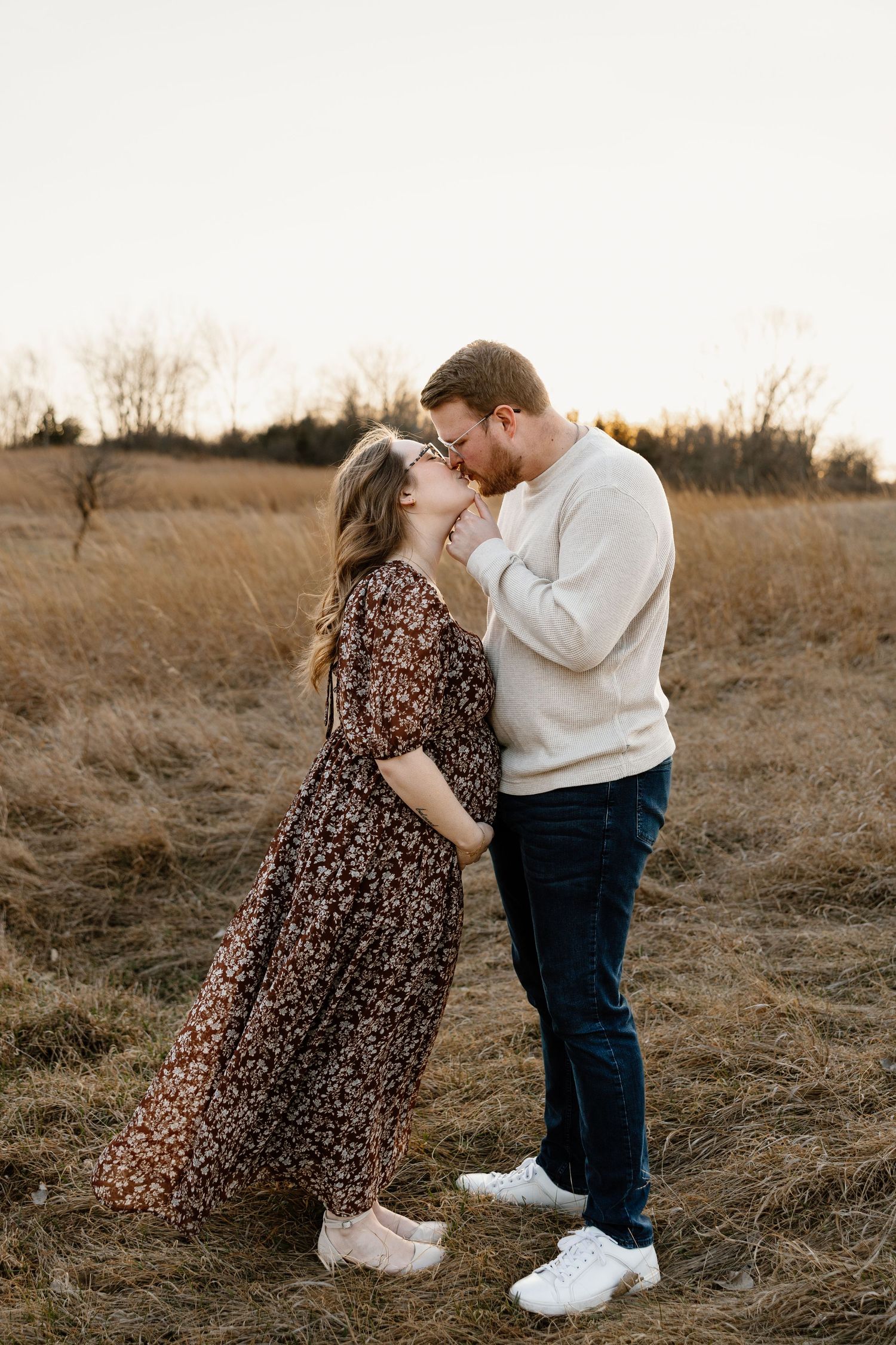 Couple sharing a romantic kiss in a golden field at sunset, woman in floral dress, man in cream sweater and jeans.