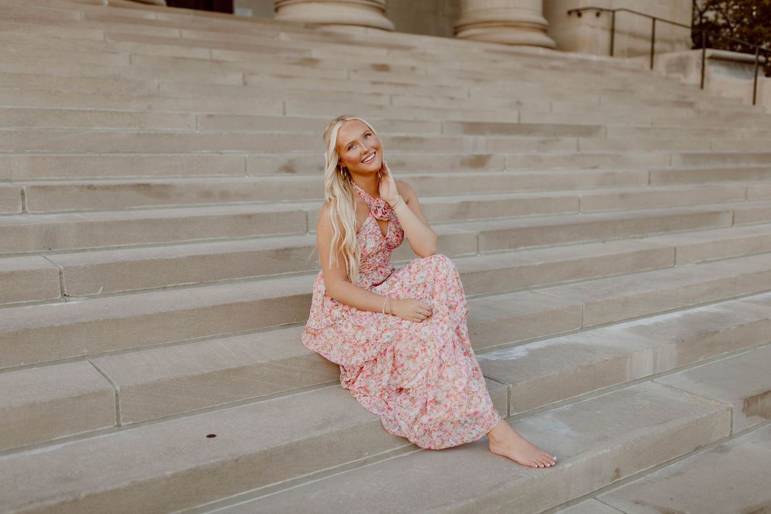 Woman in flowing pink floral dress sits gracefully on stone steps at sunset.