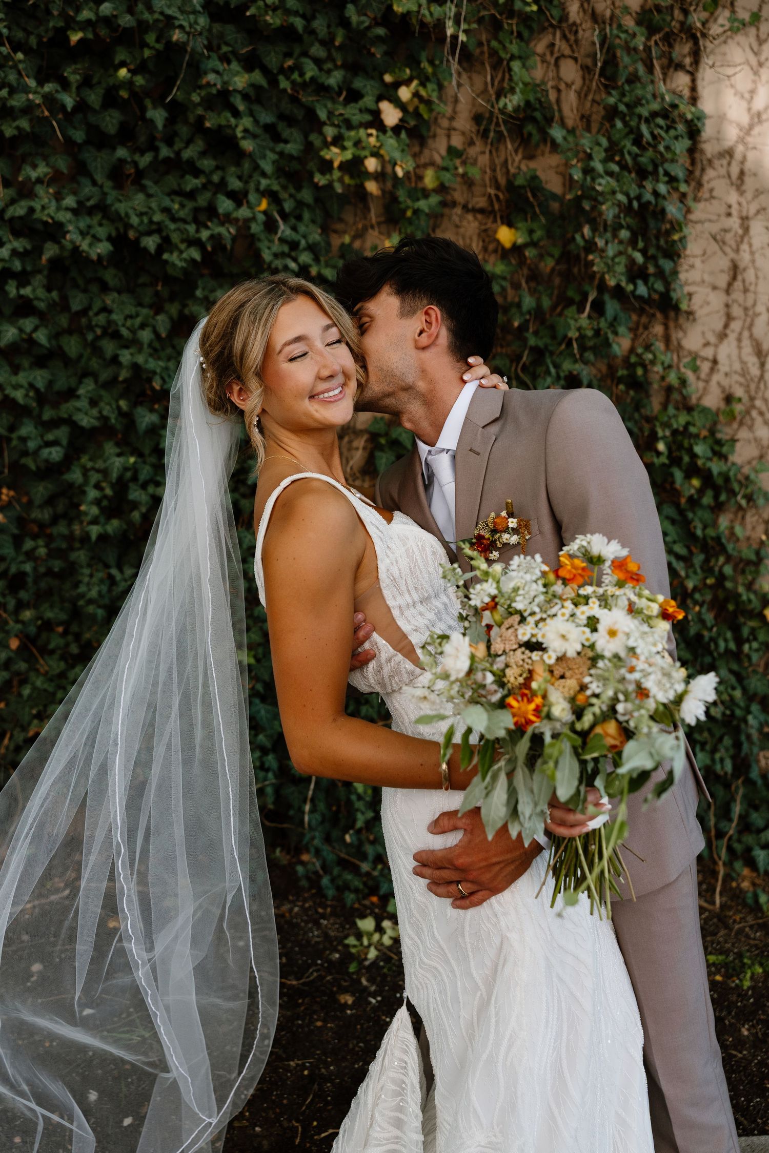 Newlyweds share tender moment with autumn-colored bouquet against ivy backdrop.