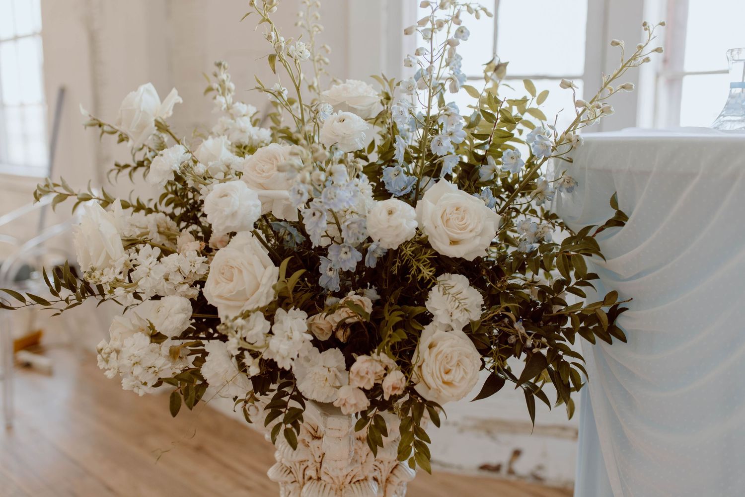 A lush white floral arrangement with roses and greenery sits elegantly in natural window light.
