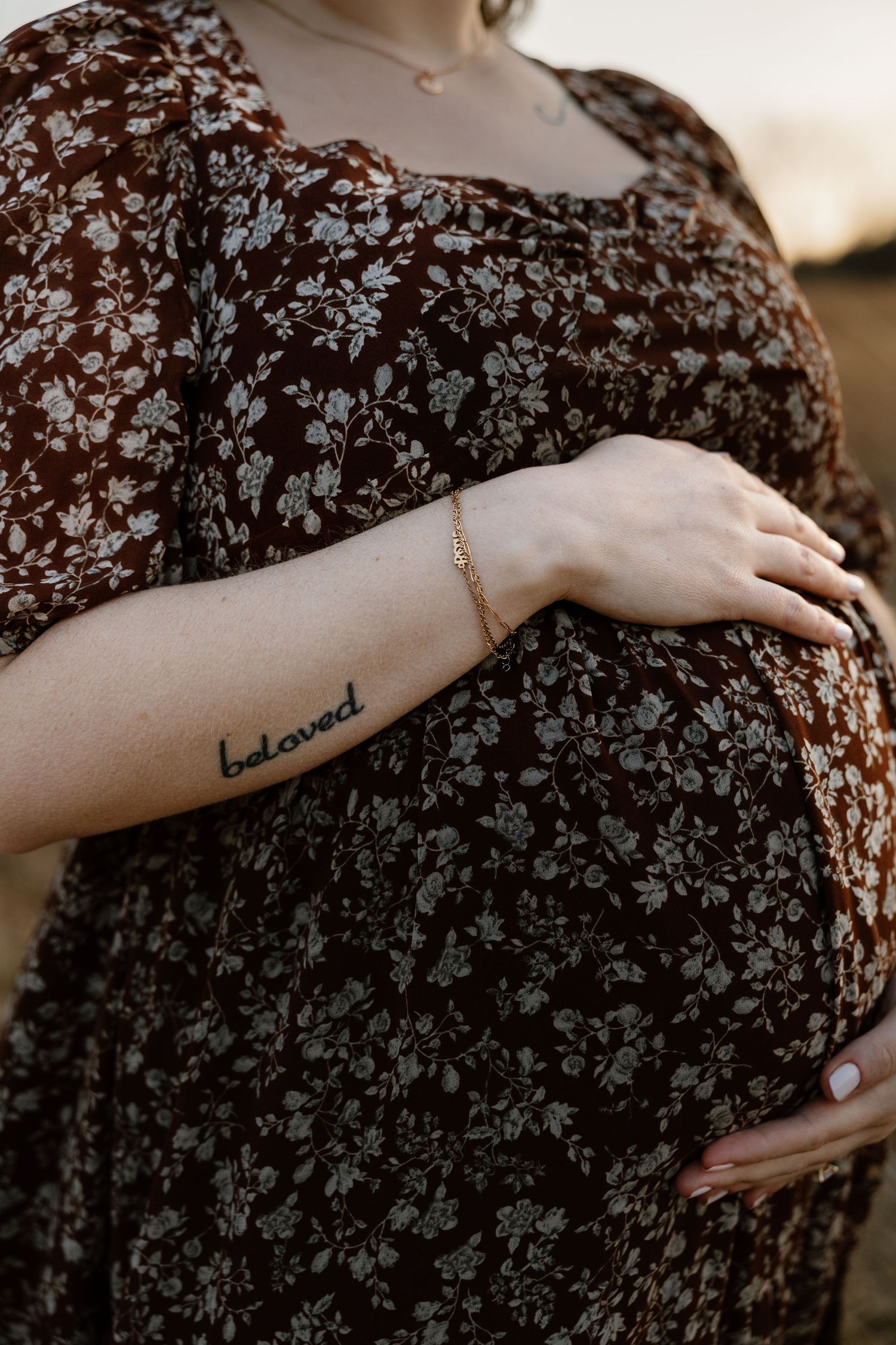 Pregnant woman in brown floral dress cradling baby bump, with a beloved tattoo and gold bracelet visible on her forearm.