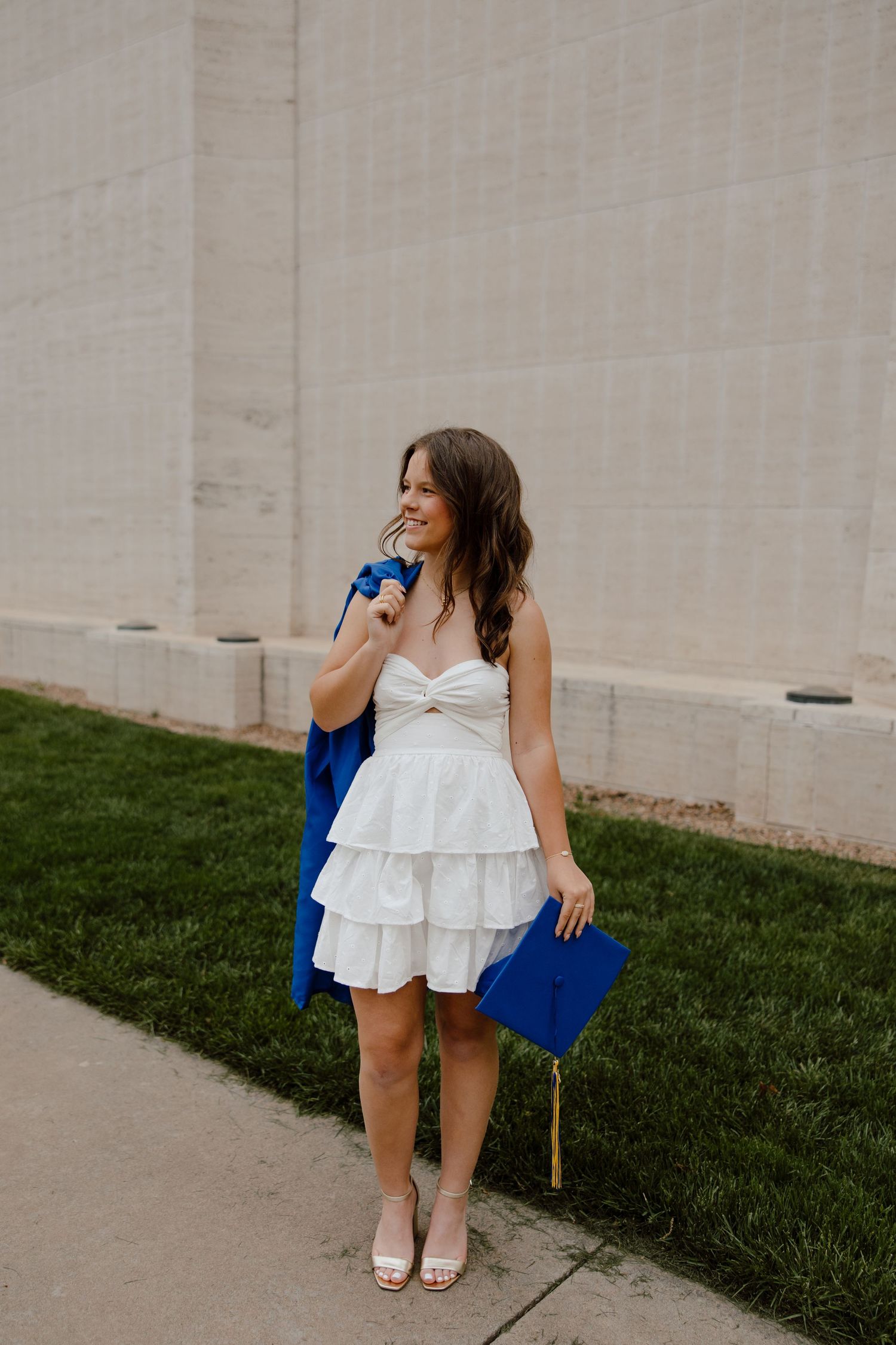 Graduate in white dress posing playfully with graduation cap on grass.
