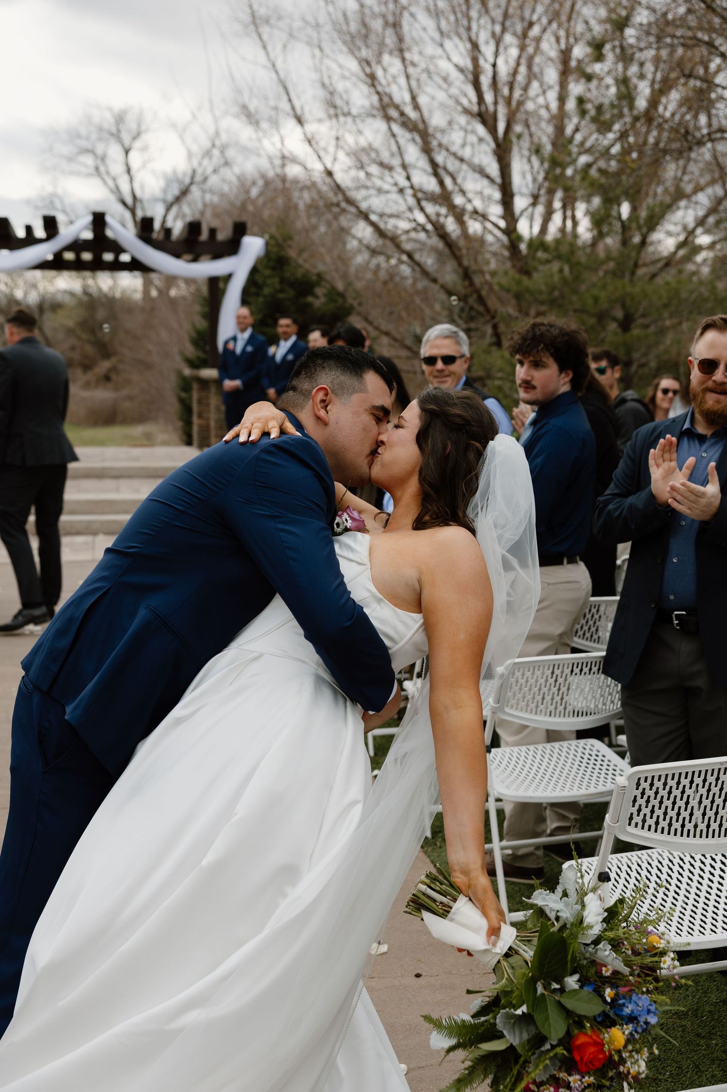 Bride and groom share first kiss at outdoor wedding ceremony with guests applauding in background.