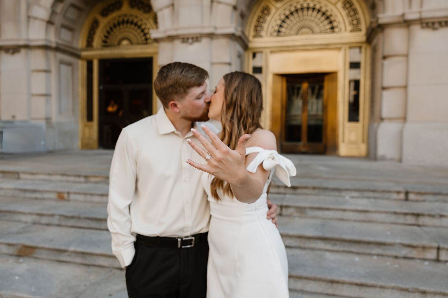 Couple kissing on steps of ornate building, bride showing engagement ring in white dress, groom in white shirt.